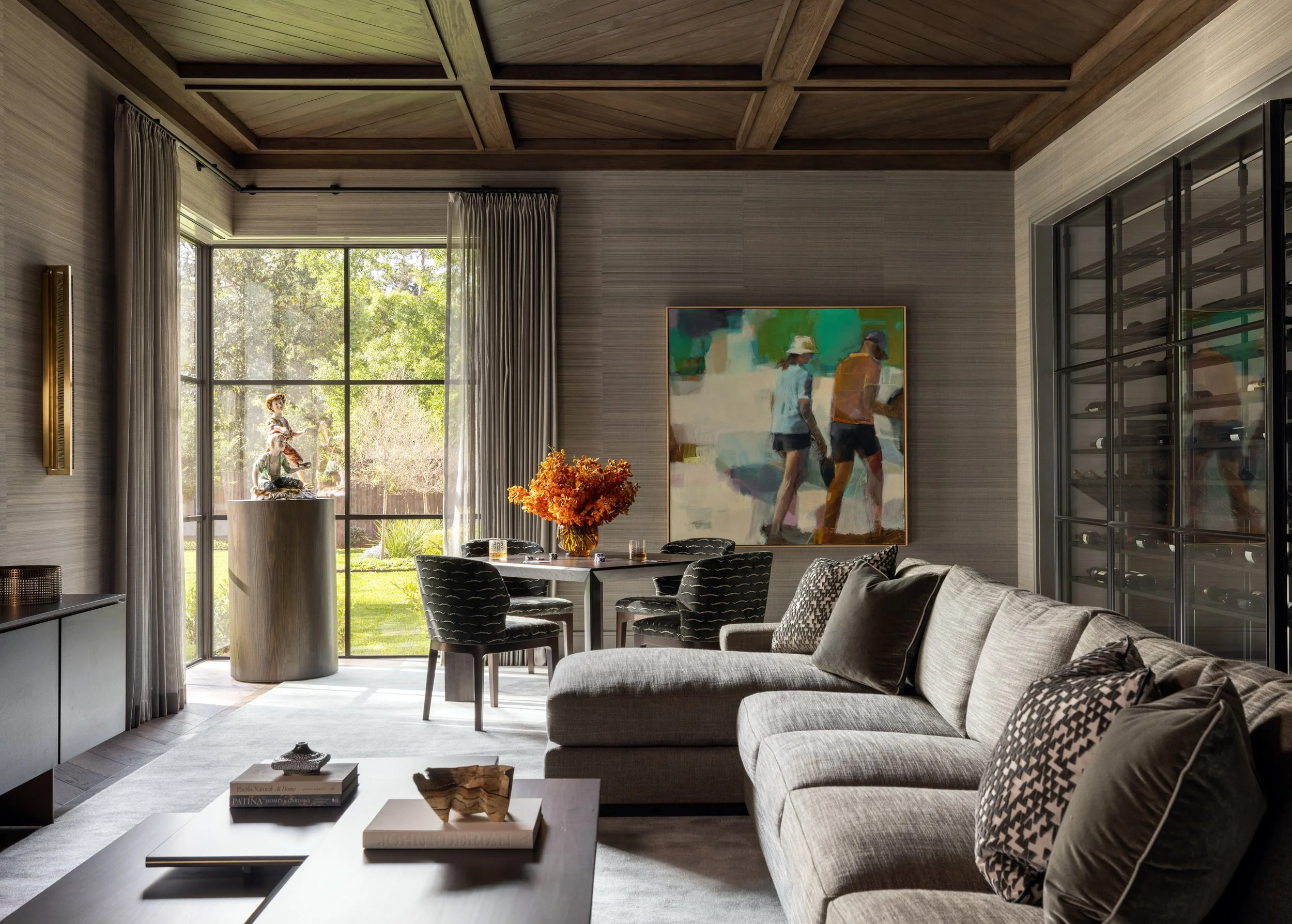 Benjamin Johnston Design Houston living room featuring coffered wood ceiling, grasscloth walls, gray sectional sofa, figurative abstract artwork, floor-to-ceiling steel windows, and built-in wine storage.