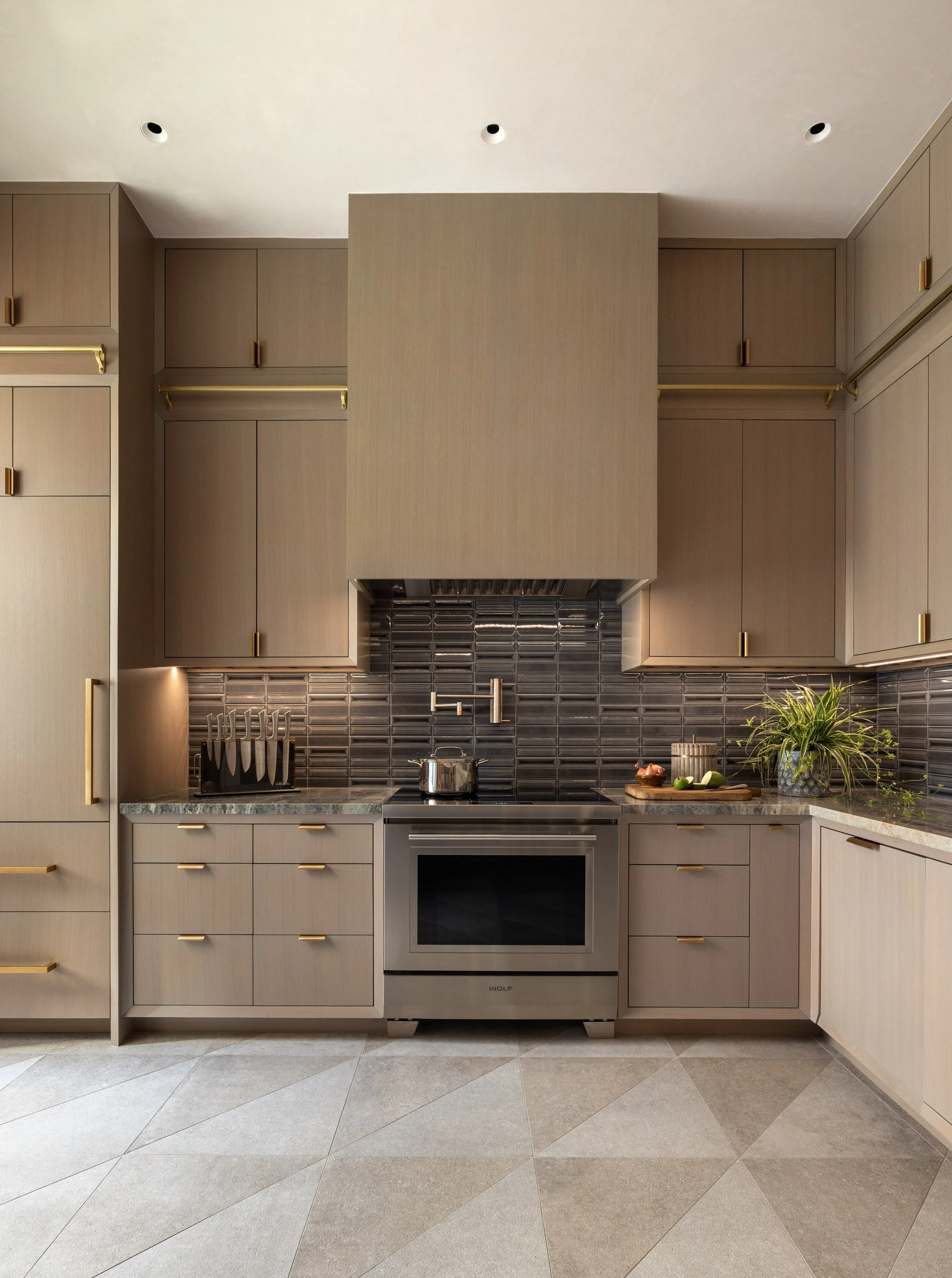 Benjamin Johnston Design Houston catering kitchen featuring taupe flat front cabinetry with brass hardware, sculptural range hood, dark glazed tile backsplash, Wolf range, and geometric stone tile floor.