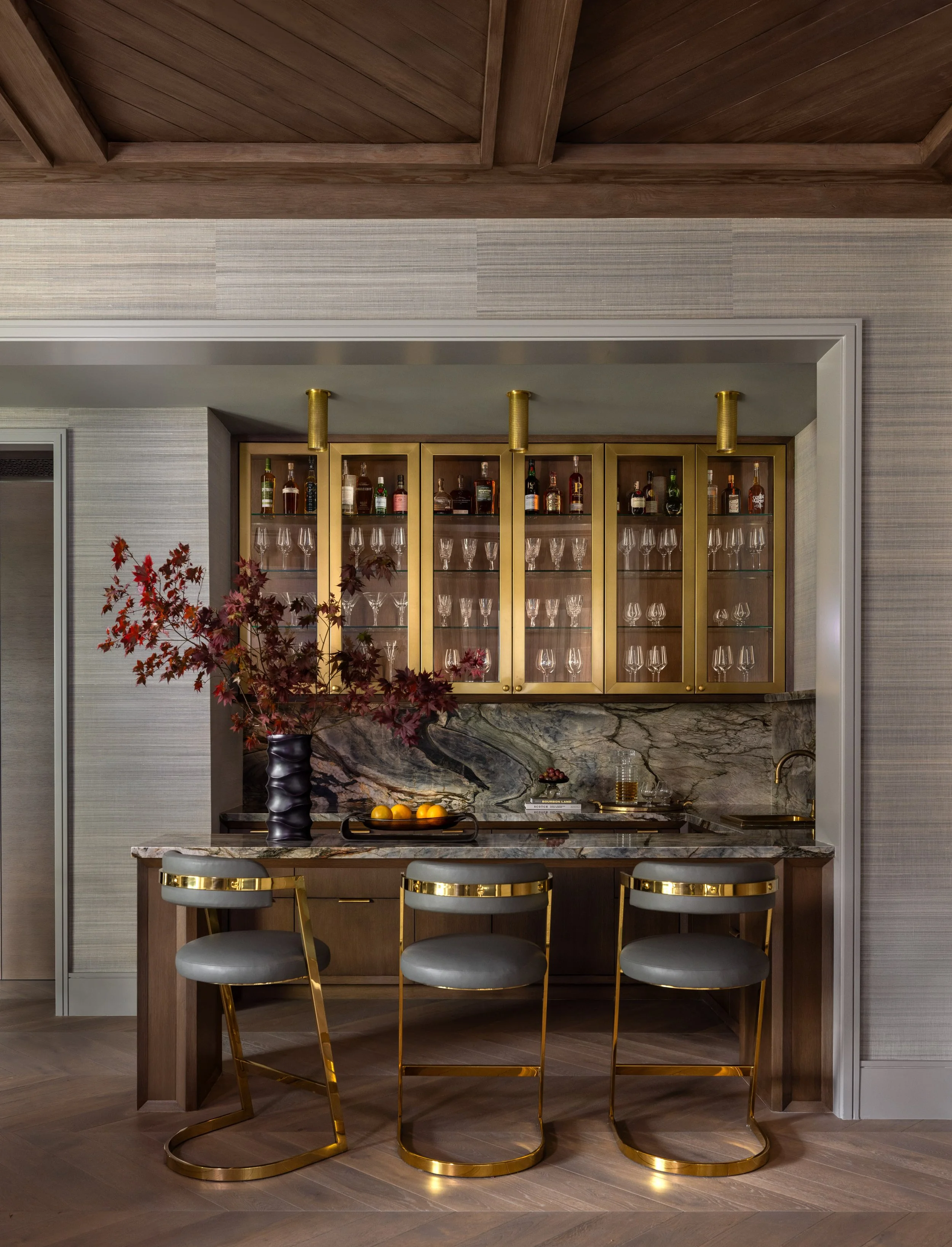 Benjamin Johnston Design Houston home bar featuring brass glass front cabinets, dramatic veined marble countertop and backsplash, gray and gold leather bar stools, and coffered wood ceiling.