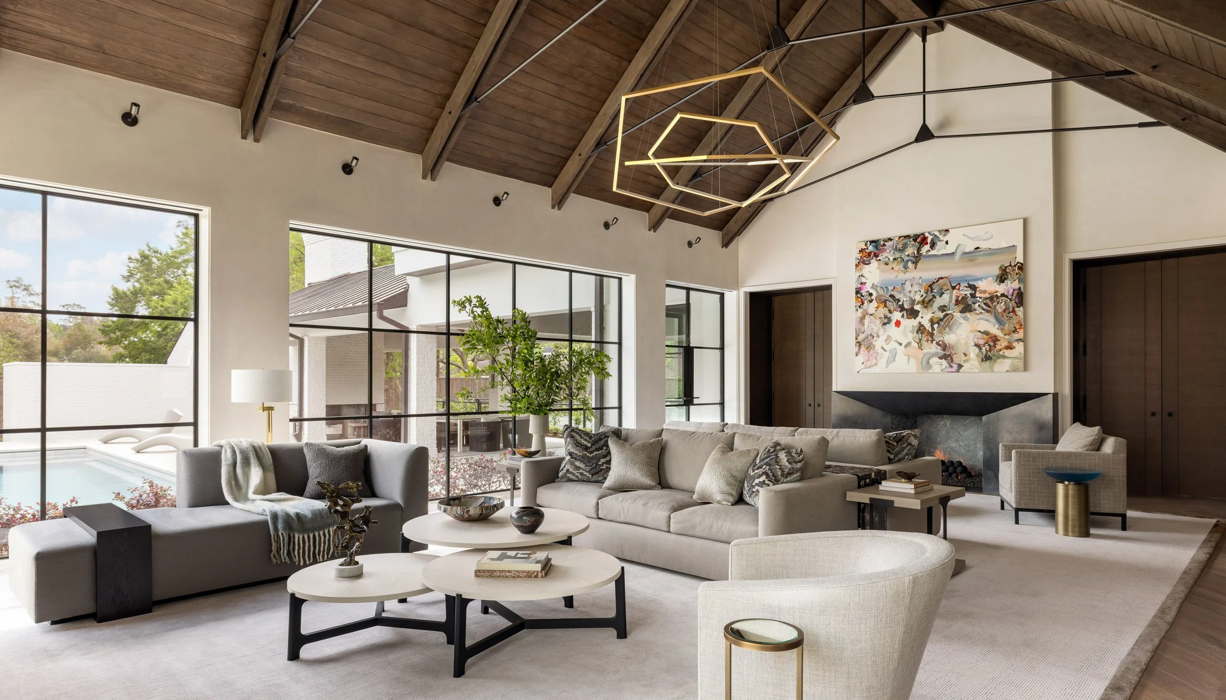 Benjamin Johnston Design great room featuring soaring vaulted wood ceiling with exposed beams, geometric brass chandelier, floor-to-ceiling steel windows overlooking pool, neutral sectional sofas, tiered round coffee tables, large scale abstract art.