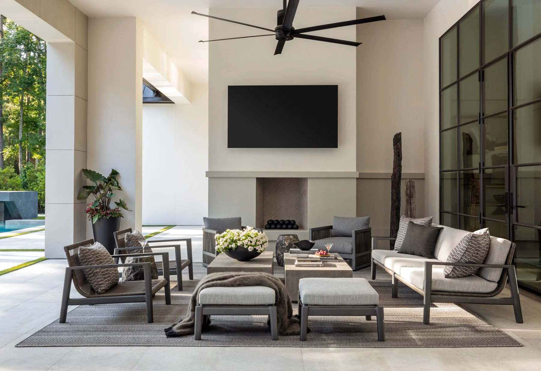 Benjamin Johnston Design Houston luxury residential interior design — covered outdoor living area with limestone fireplace, mounted TV, teak and woven lounge seating, concrete coffee table, steel frame doors, and pool view.