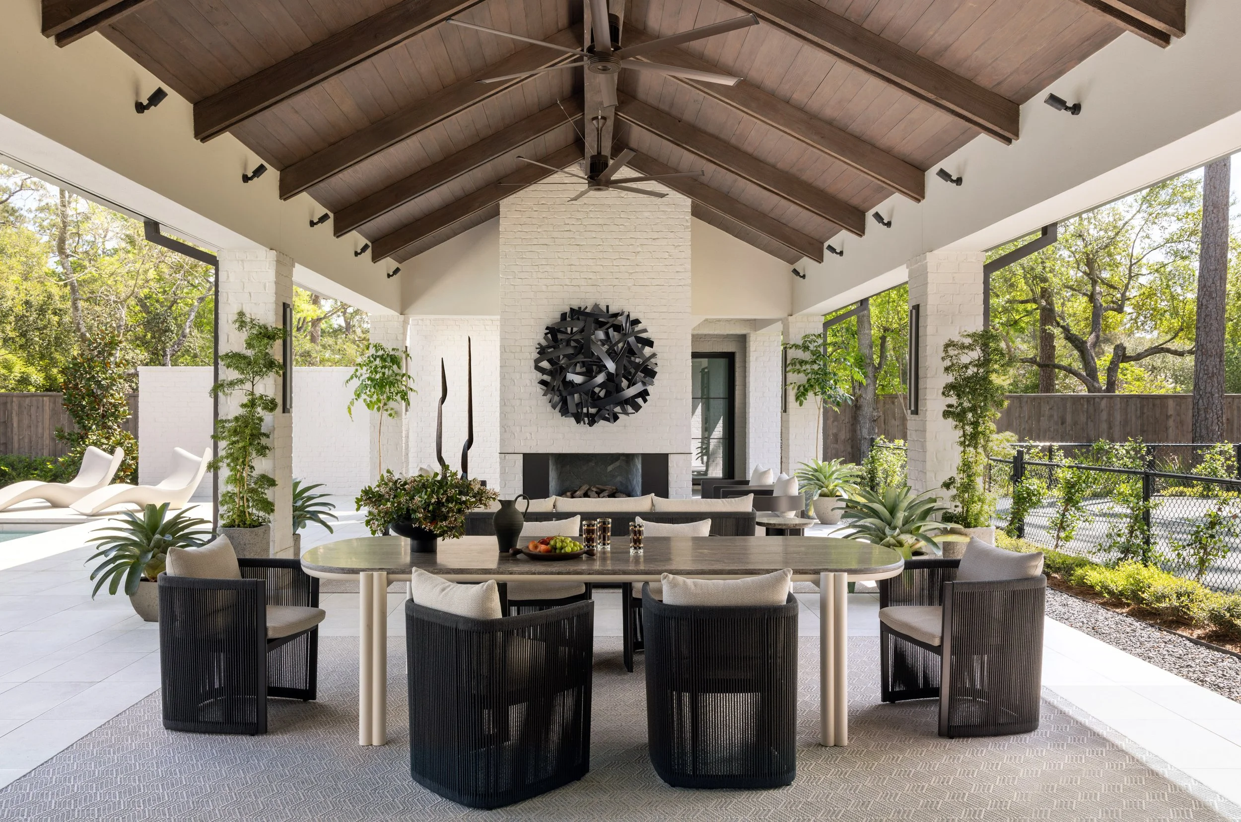 Benjamin Johnston Design Houston outdoor pavilion featuring vaulted wood ceiling with exposed beams, white brick fireplace with sculptural wall art, outdoor dining table with woven chairs, and pool view surrounded by lush landscaping.