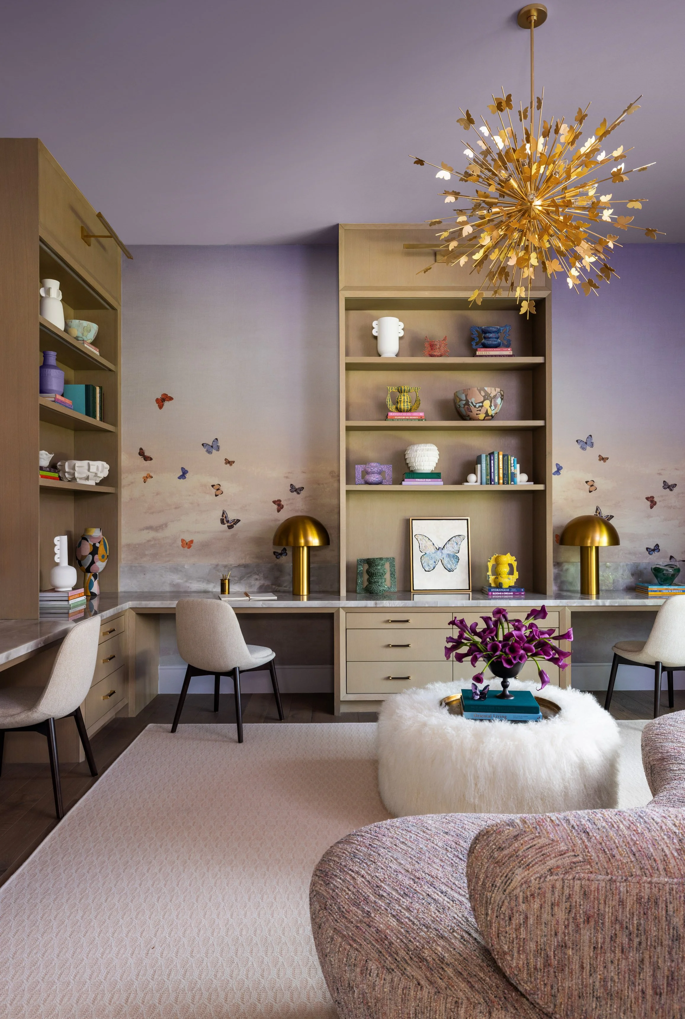A stylish living room with built-in bookshelves filled with colorful books and sculptures, a marble countertop desk with beige chairs, a large beige rug, a plush white fur ottoman with a purple orchid on top, and a gold starburst chandelier hanging f