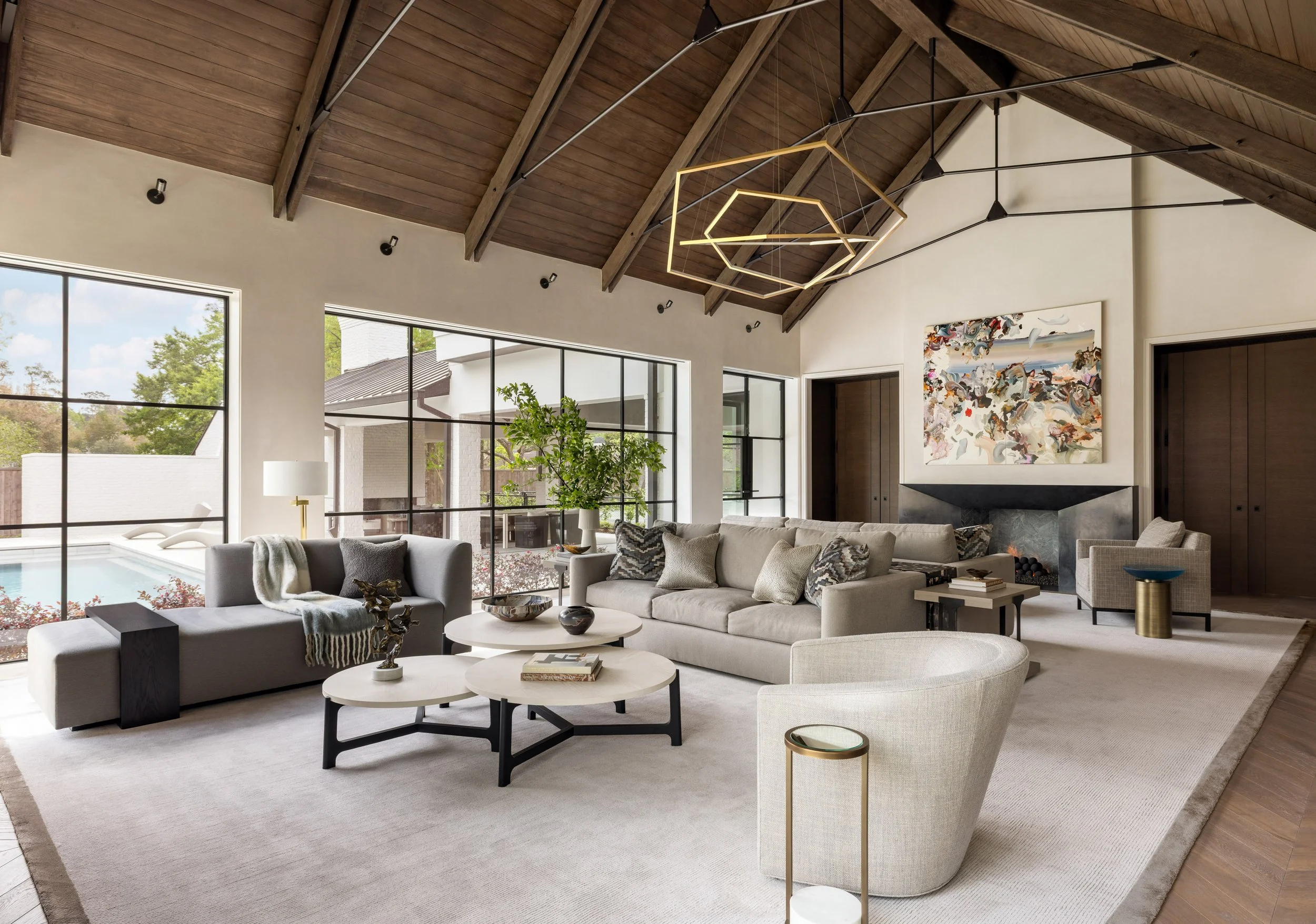 Benjamin Johnston Design living room featuring vaulted wood ceiling with exposed beams, geometric brass chandelier, floor-to-ceiling steel windows overlooking pool, neutral sectional sofas, tiered round coffee tables, abstract artwork, and stone fire