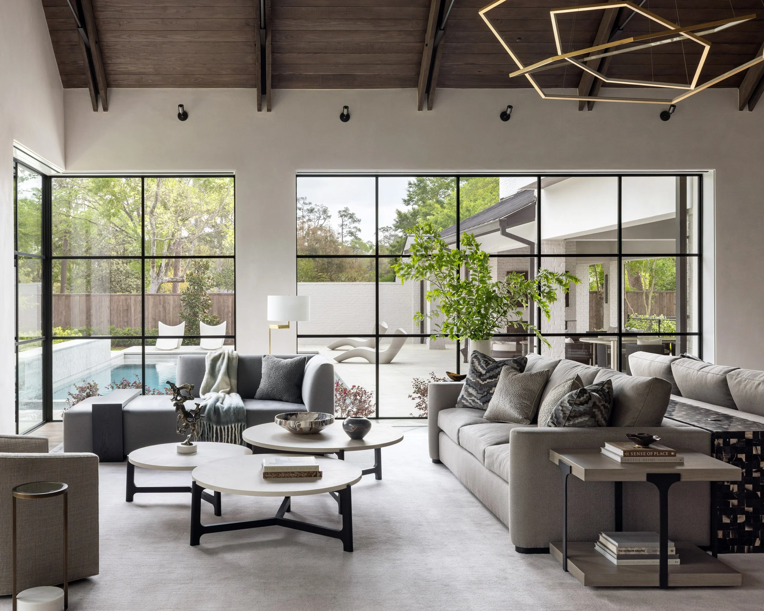 Benjamin Johnston Design living room with panoramic steel window walls framing pool and mature tree views, geometric brass chandelier, vaulted wood ceiling, gray and linen sectional sofas, tiered round coffee tables, and large indoor botanical tree.