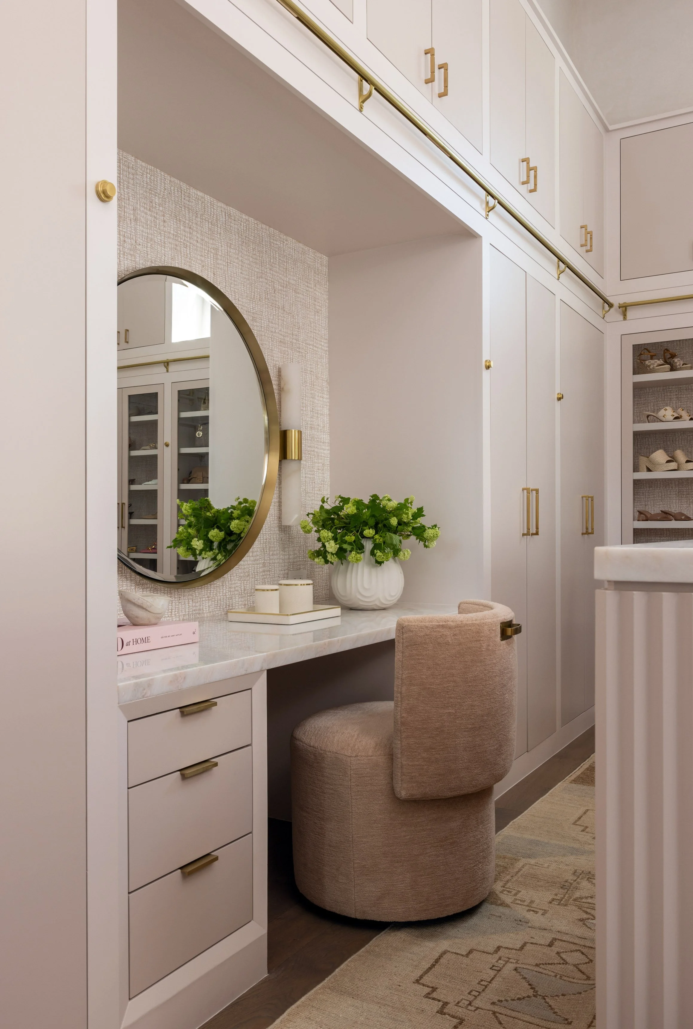 A cozy dressing area with a white built-in vanity with six drawers and gold handles, topped with a white marble surface. A large round mirror with a gold frame hangs above the vanity. On the vanity, there are a white potted plant with green foliage, 