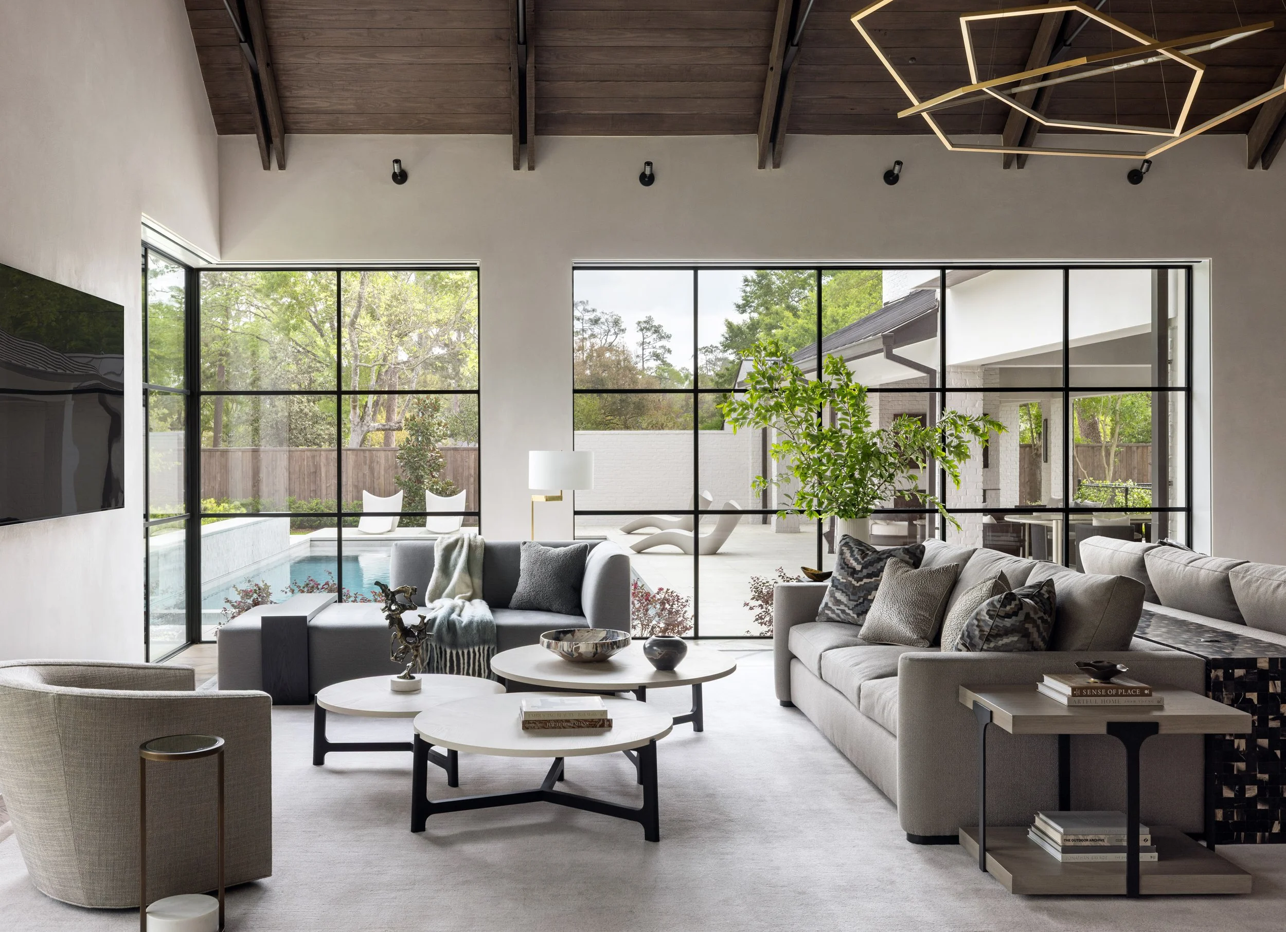 Benjamin Johnston Design living room featuring floor-to-ceiling steel windows with pool and courtyard views, geometric brass chandelier, vaulted wood ceiling, neutral sectional sofas, tiered round coffee tables, and curved lounge chair.