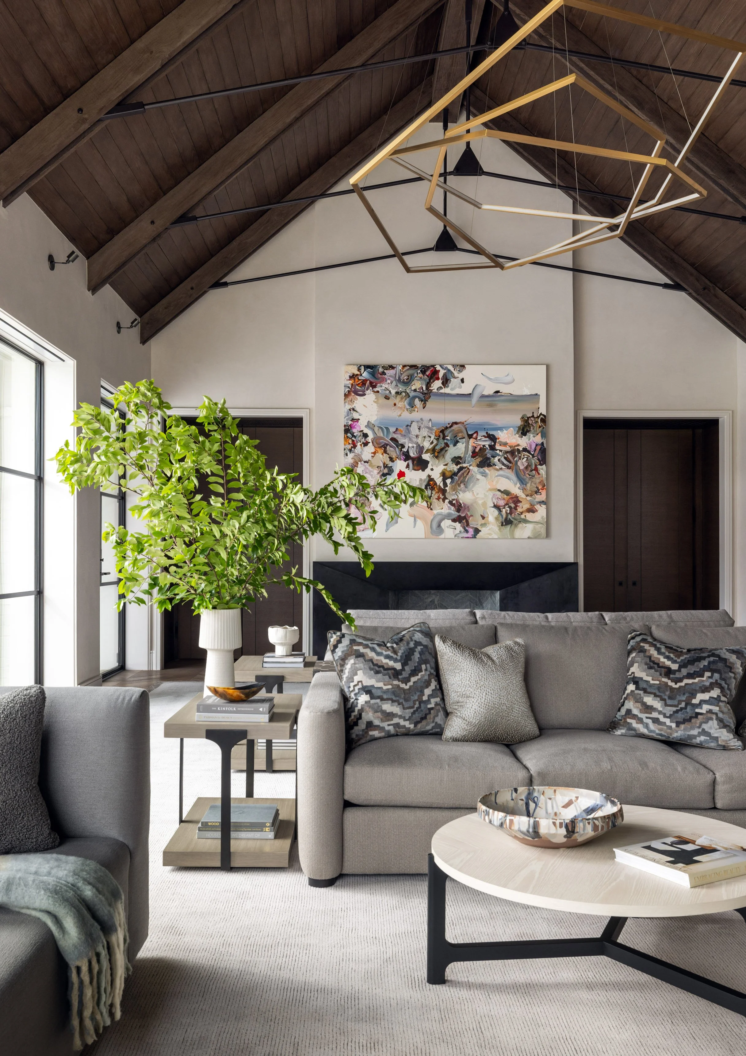 Benjamin Johnston Design living room featuring vaulted wood ceiling with exposed beams, geometric brass chandelier, gray sectional sofa with chevron patterned pillows, round stone coffee table, and abstract artwork above stone fireplace.