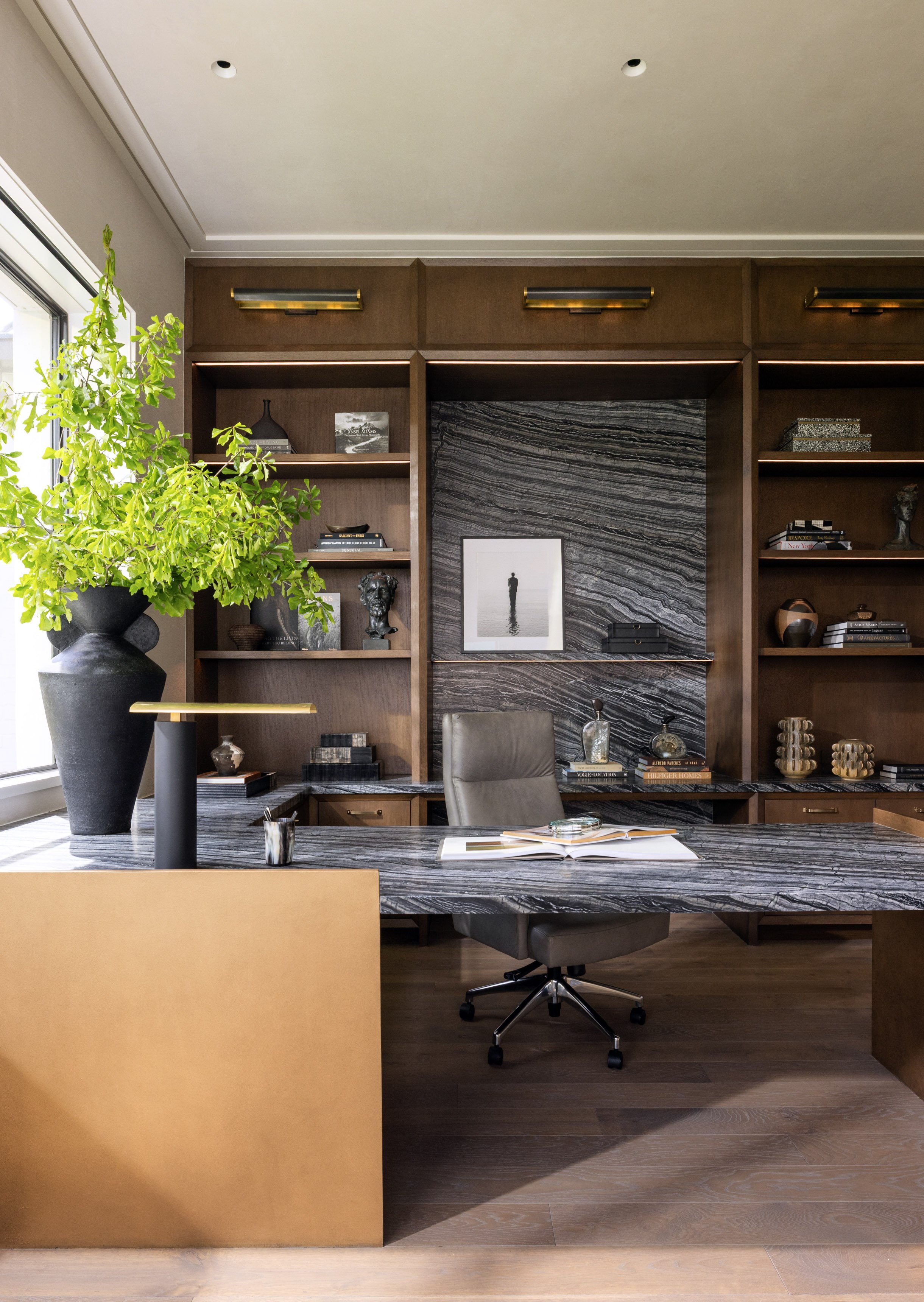 Benjamin Johnston Design home office with veined marble desk and accent wall, full wall walnut built-in shelving with brass picture lights, leather desk chair, sculptural black vase with greenery, and wide plank wood floor.
