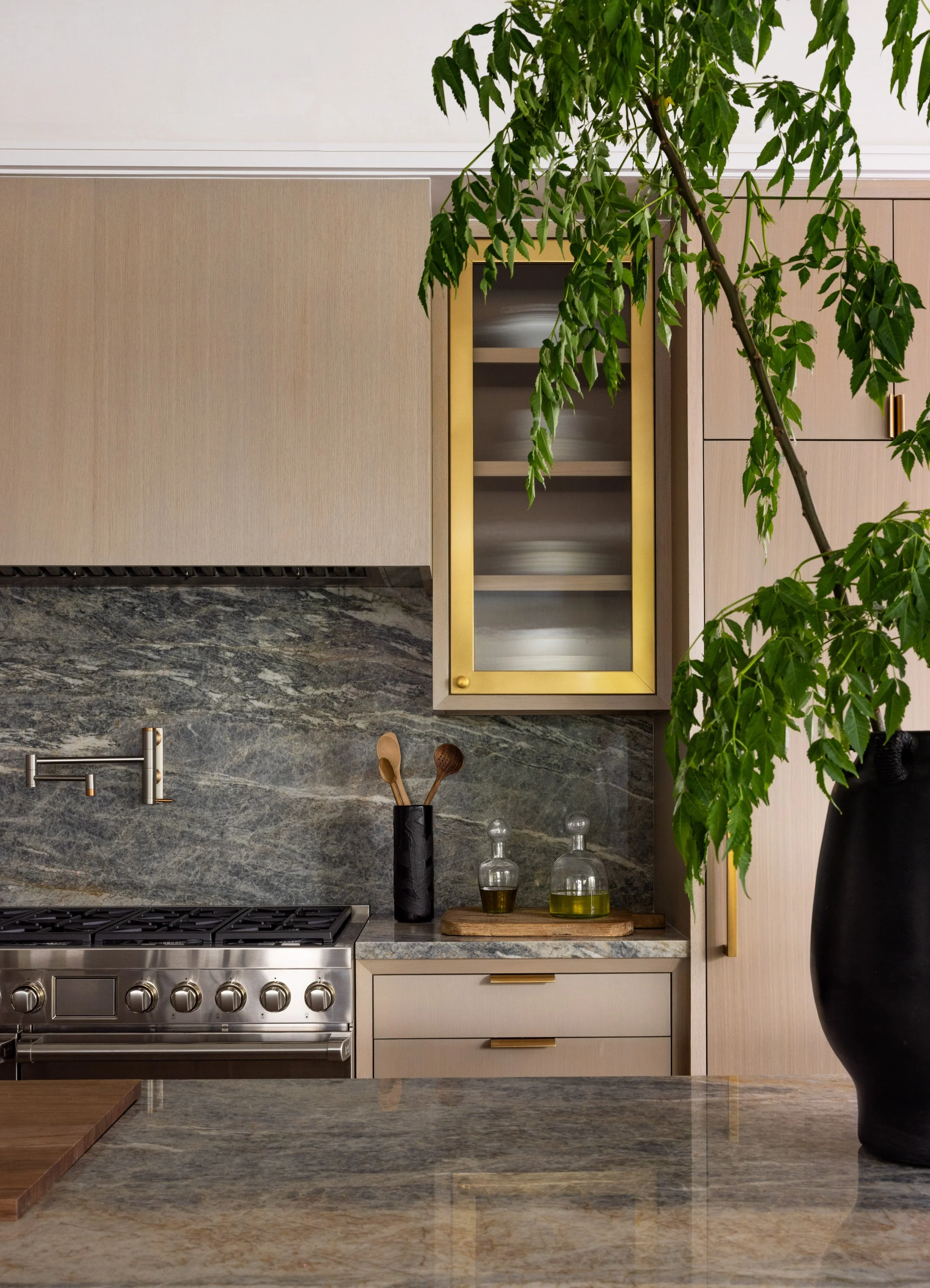 Modern kitchen with beige cabinets, stainless steel stove, dark marble backsplash, wooden cutting board, black vase with wooden spoons, glass bottles with oil or vinegar, and a large black vase with a tall green plant.