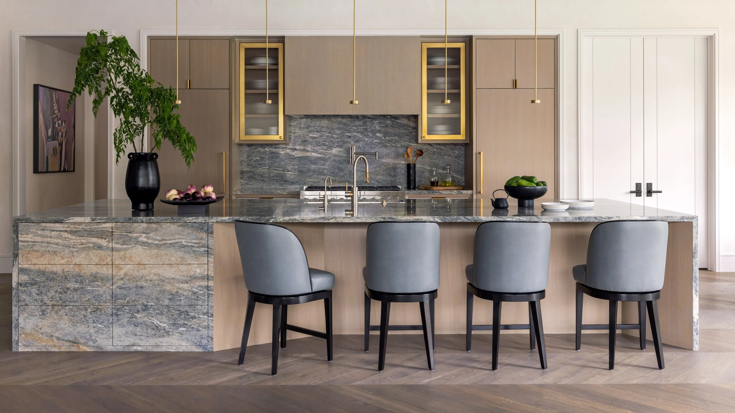 Benjamin Johnston Design Houston kitchen featuring large scale marble waterfall island, gray leather bar stools, brass framed glass upper cabinets, veined marble backsplash, and herringbone wood floor.