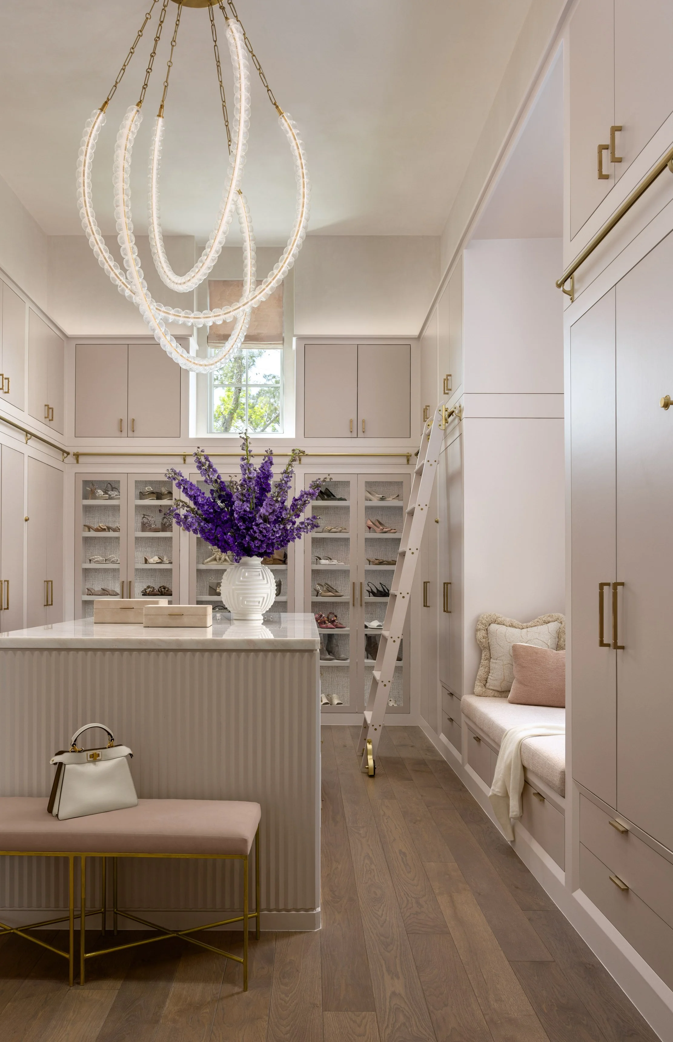 A modern walk-in closet with built-in cabinets, a large chandelier, a marble island with a purple flower arrangement, and seating with pillows. There is a small suitcase on a bench and a ladder leaning against the cabinets.