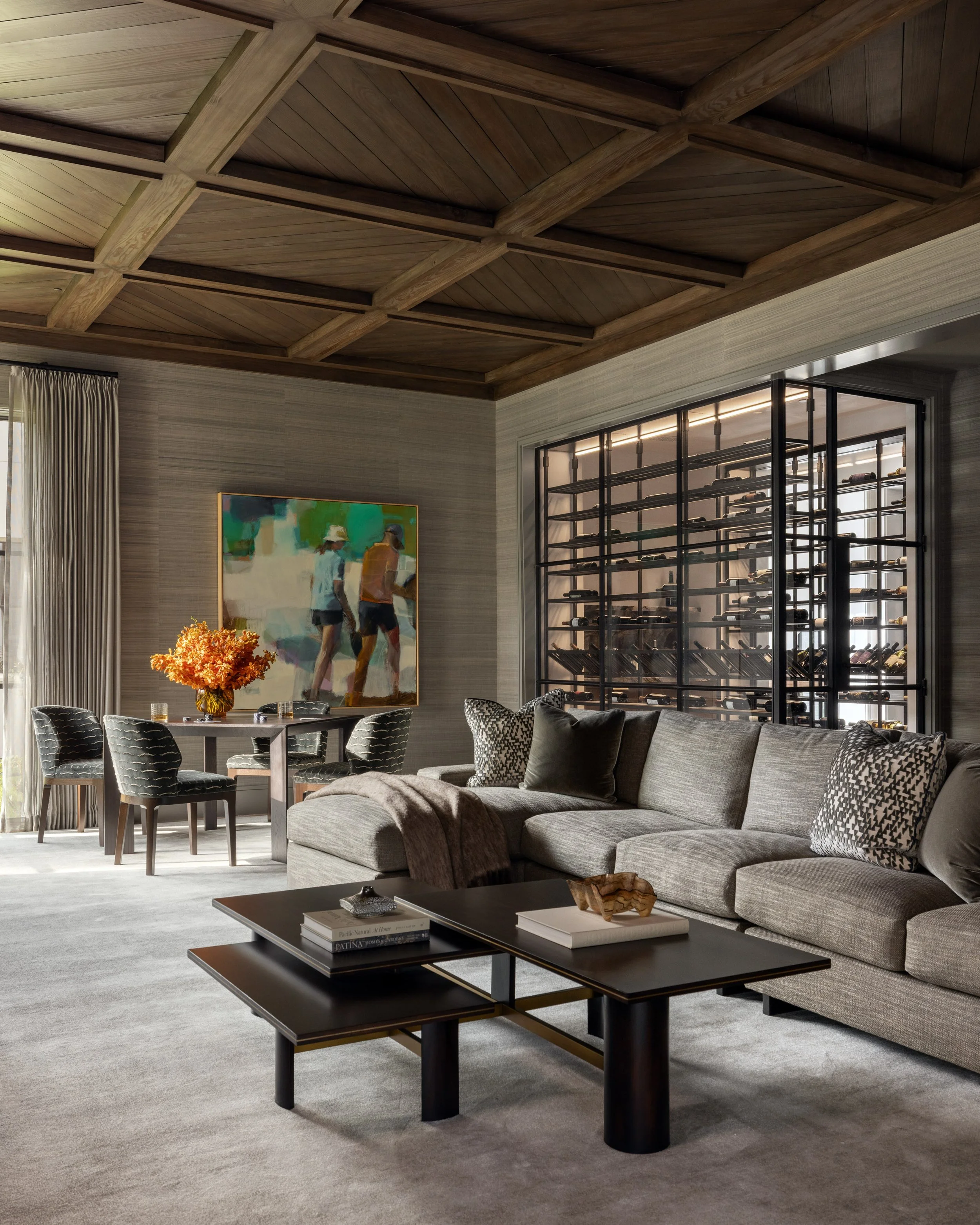 Image of a modern living room with a wood-paneled coffered ceiling, a large L-shaped gray sofa with decorative pillows and a blanket, a dark coffee table with books and decorative objects, a round dining table with velvet upholstered chairs, a large 