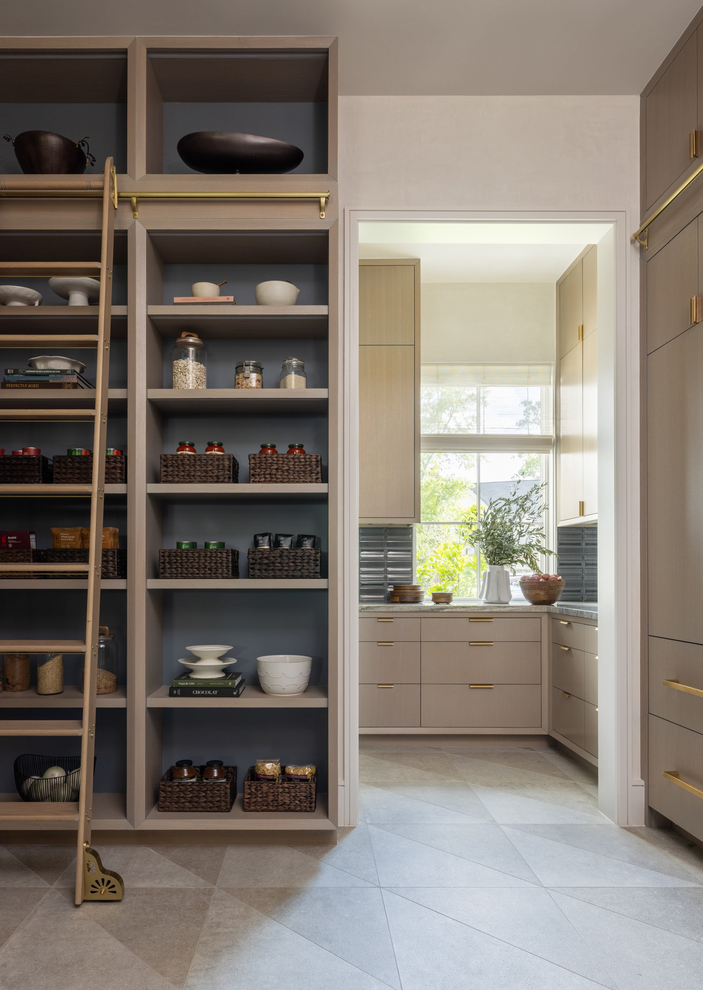 A view of a modern kitchen with a built-in wooden shelving unit on the left filled with jars, bowls, books, and baskets. To the right is a doorway leading into a bright room with a window, a plant, and kitchen cabinets with gold handles.