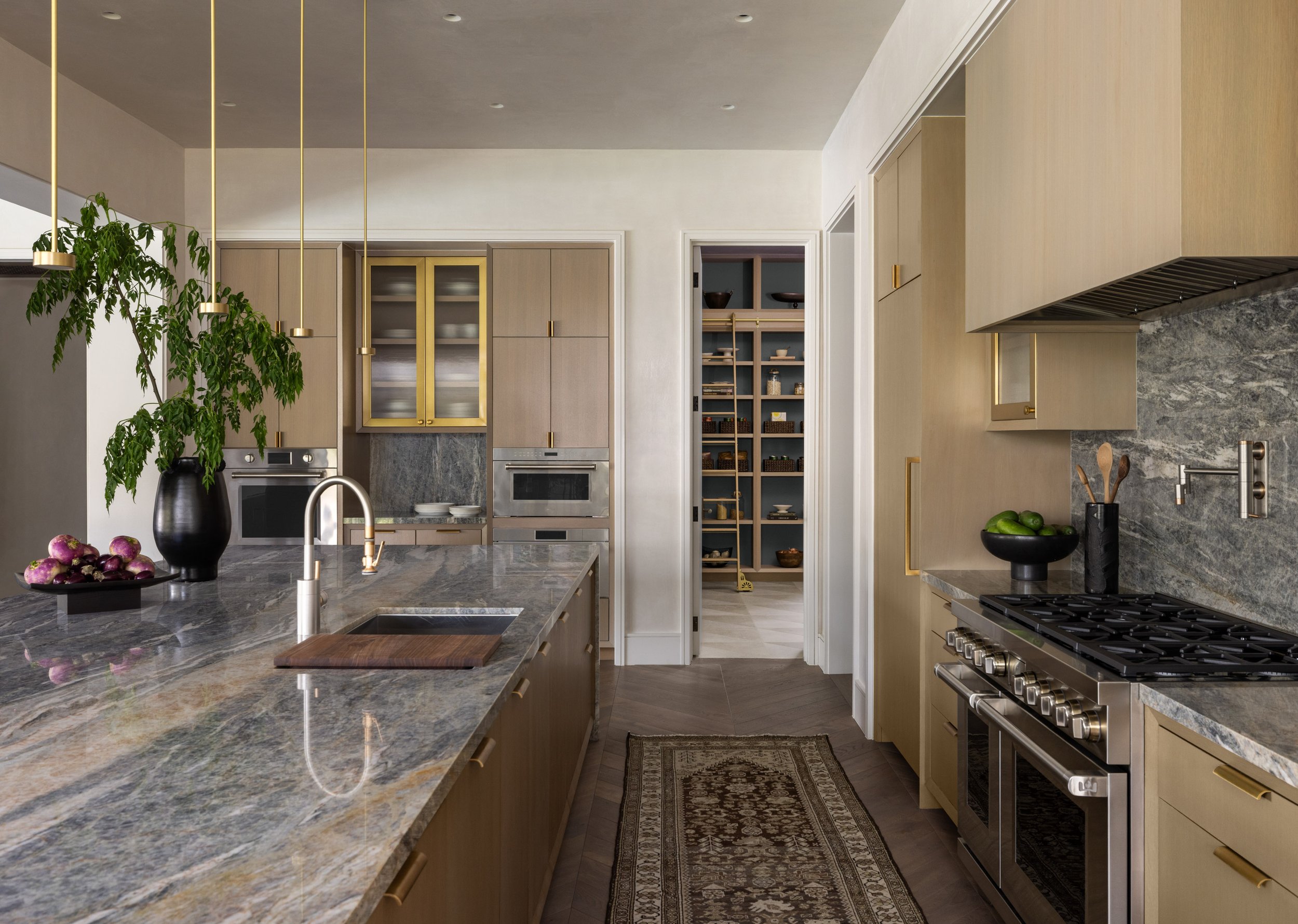 Benjamin Johnston Design Houston kitchen featuring veined marble countertops and backsplash, flat front cabinetry with brass hardware, professional range, brass pendant lights, vintage runner rug, and open pantry with library ladder.