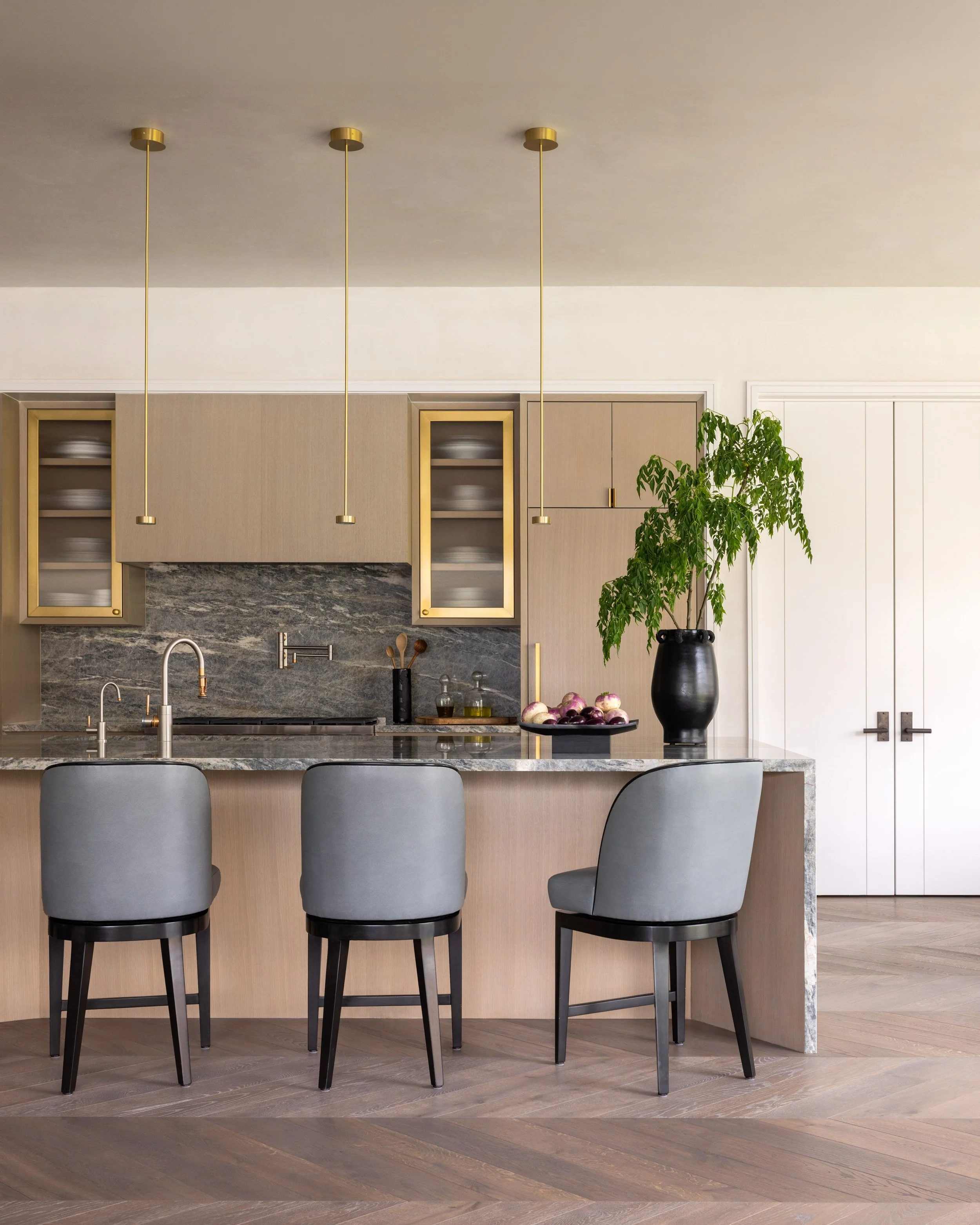 Modern kitchen with a marble island, three gray bar stools, a black vase with a leafy green plant, and gold pendant lights hanging from the ceiling.