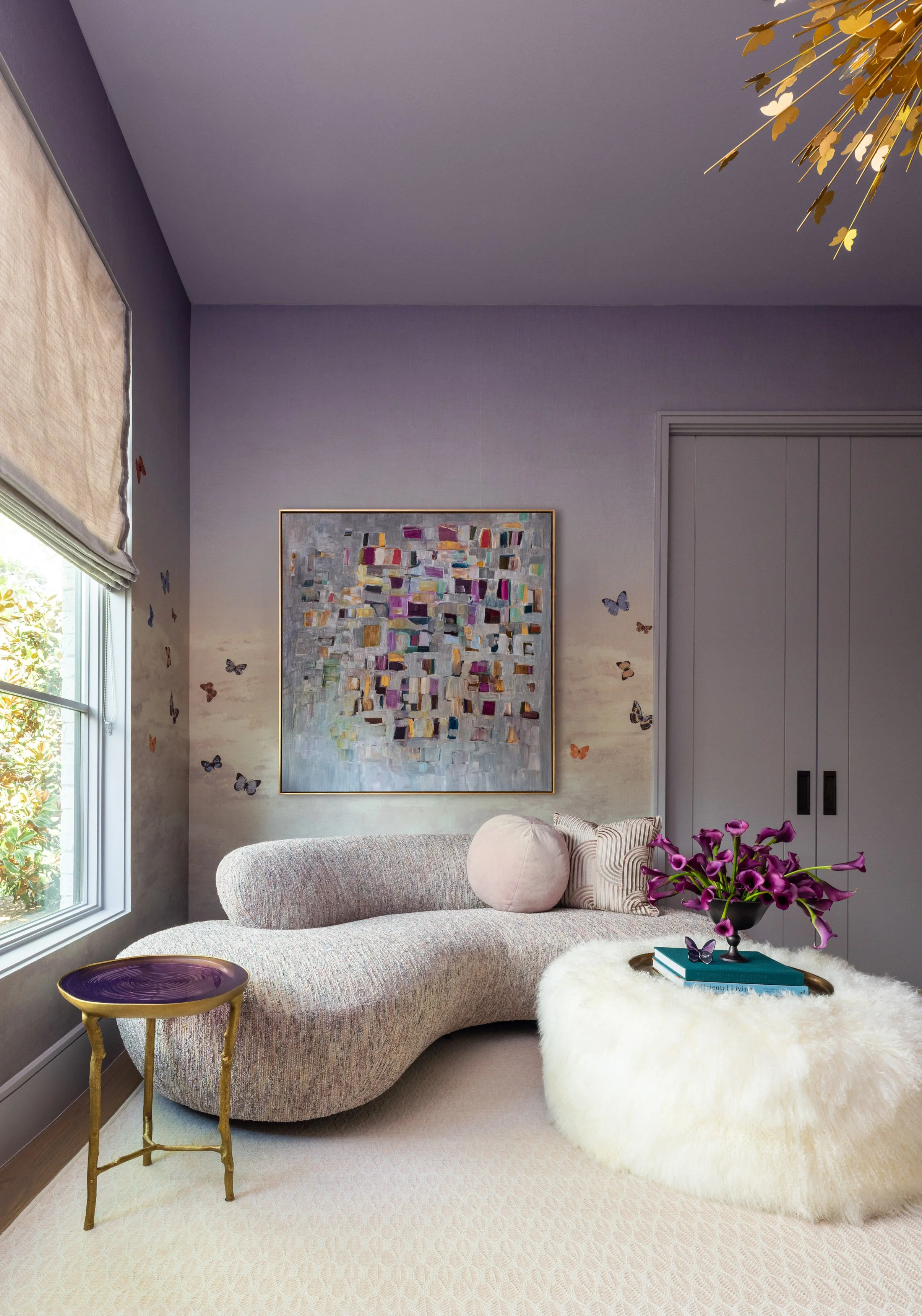 A living room with a beige curved sofa, a patterned pillow, a fluffy white ottoman with a pink pillow, a vase of purple flowers, a tray with books, a colorful abstract artwork on the wall, butterflies on the wall, a window with a beige shade, and a g
