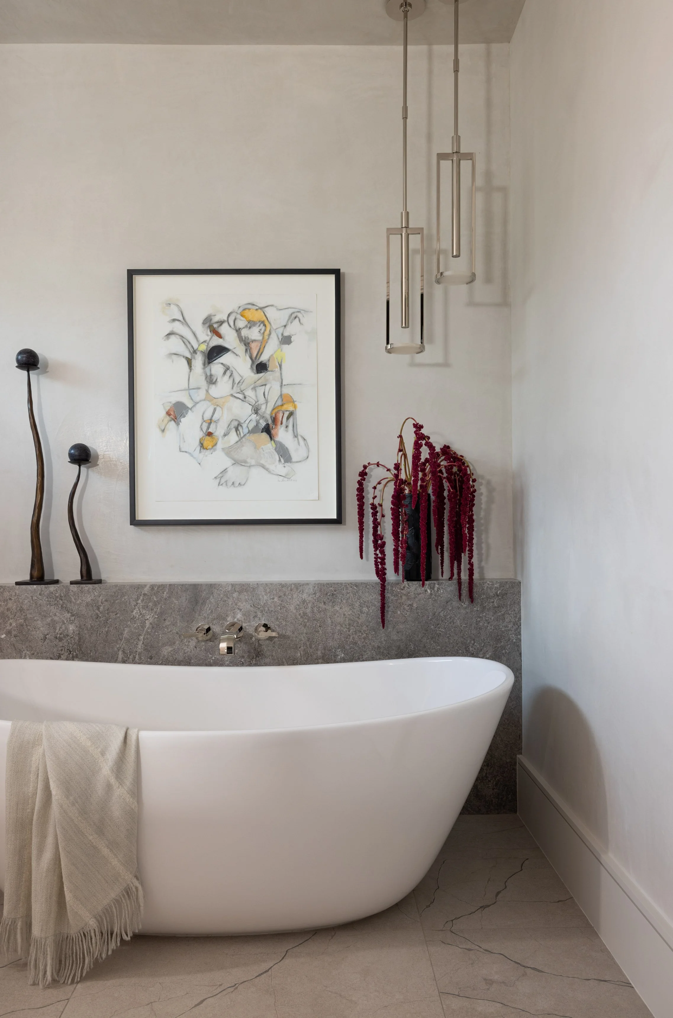 Modern bathroom with a white freestanding bathtub, beige towel, framed abstract art on the wall, dark wooden sculptures, and a red flower arrangement with hanging blooms. There are three contemporary pendant lights hanging from the ceiling and marble