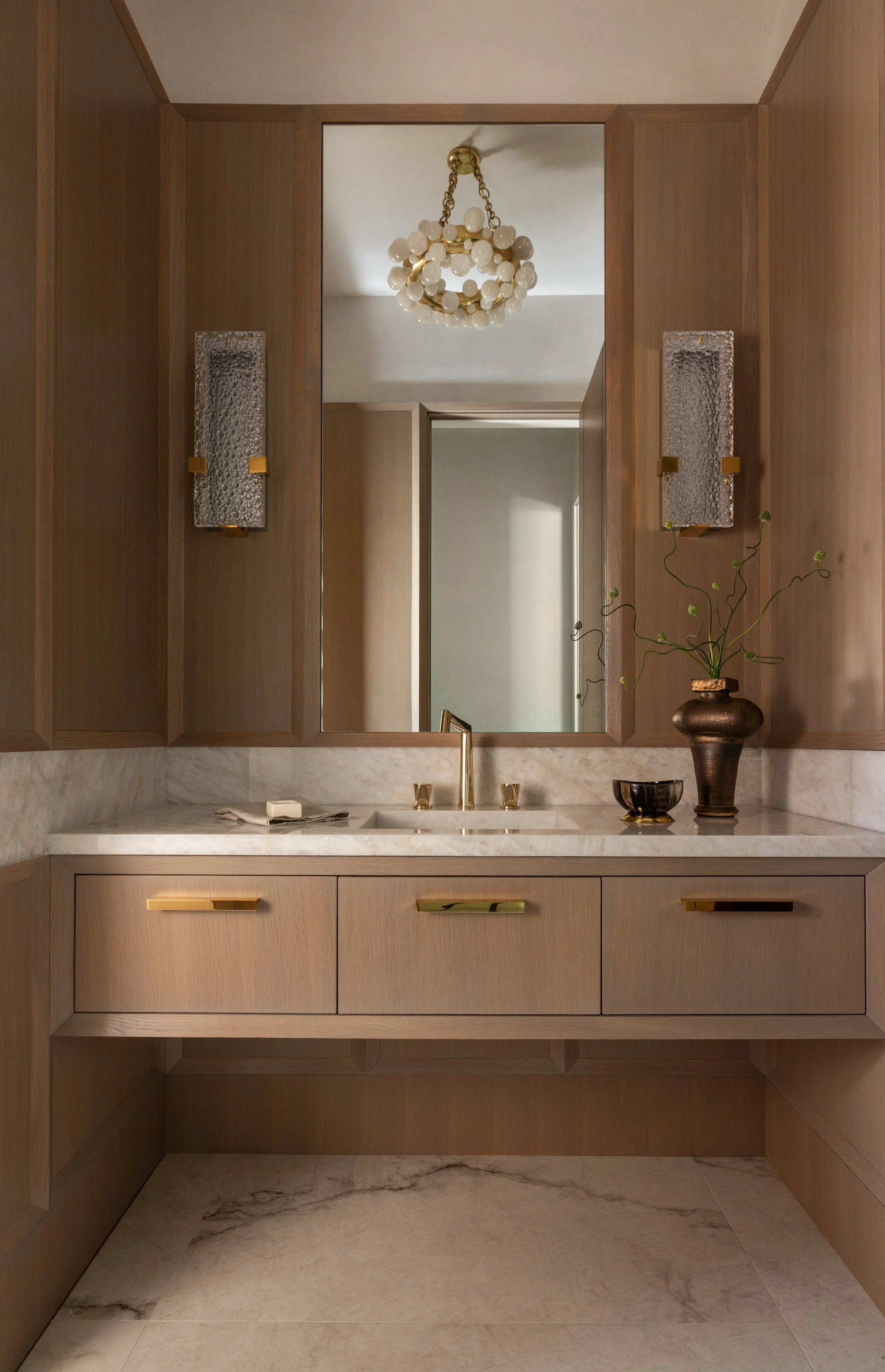A bathroom vanity with a marble countertop, a gold faucet, a decorative bowl, and a brown vase with green stemmed plants. Behind the vanity, a large mirror is flanked by two textured wall sconces with gold accents. Above, a chandelier with multiple w
