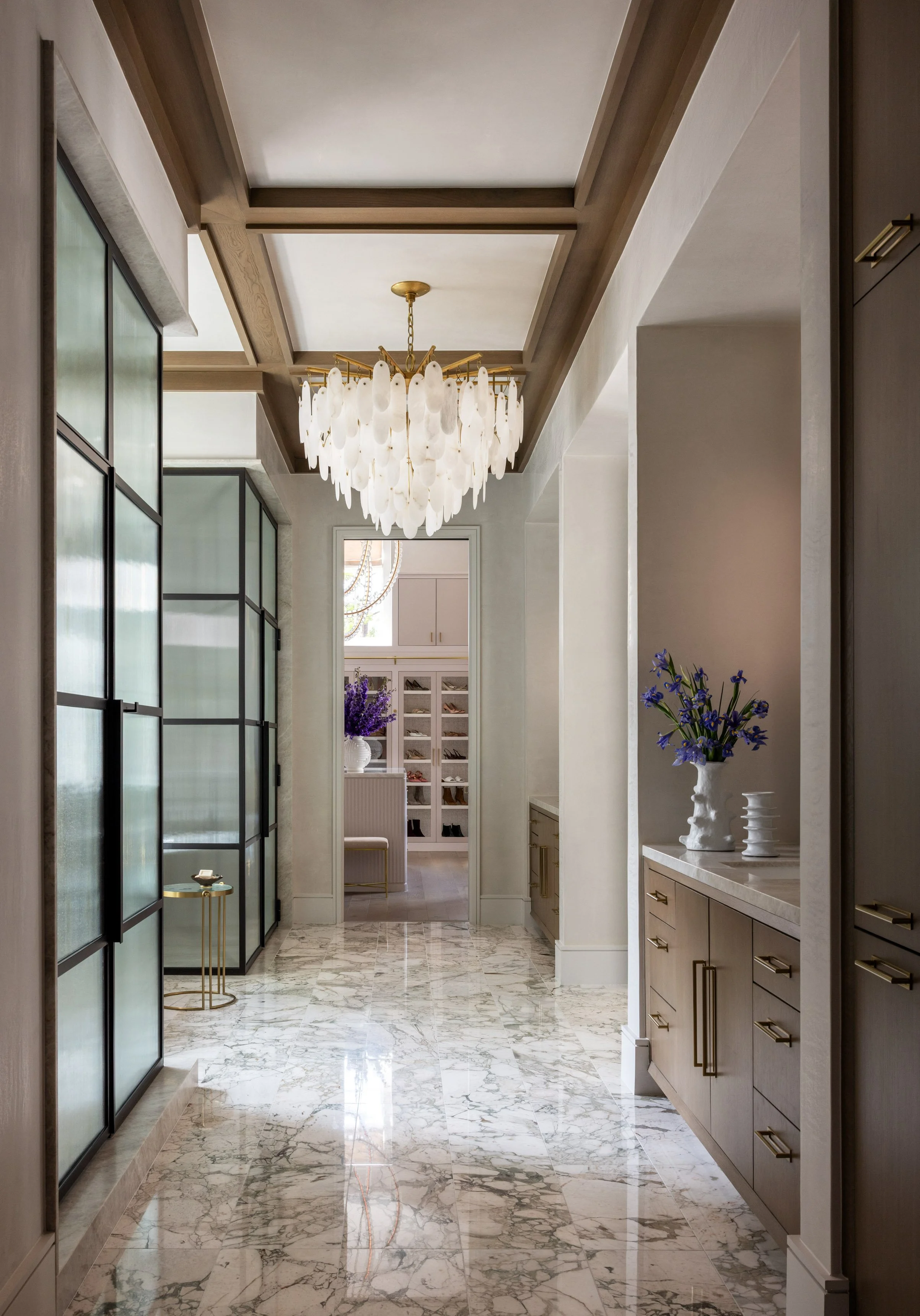 Elegant hallway with marble flooring, built-in cabinets, and a chandelier hanging from the ceiling. There are large frosted glass doors on the left and decorative vases with purple flowers on the right. At the end, a door opens to a room with white c