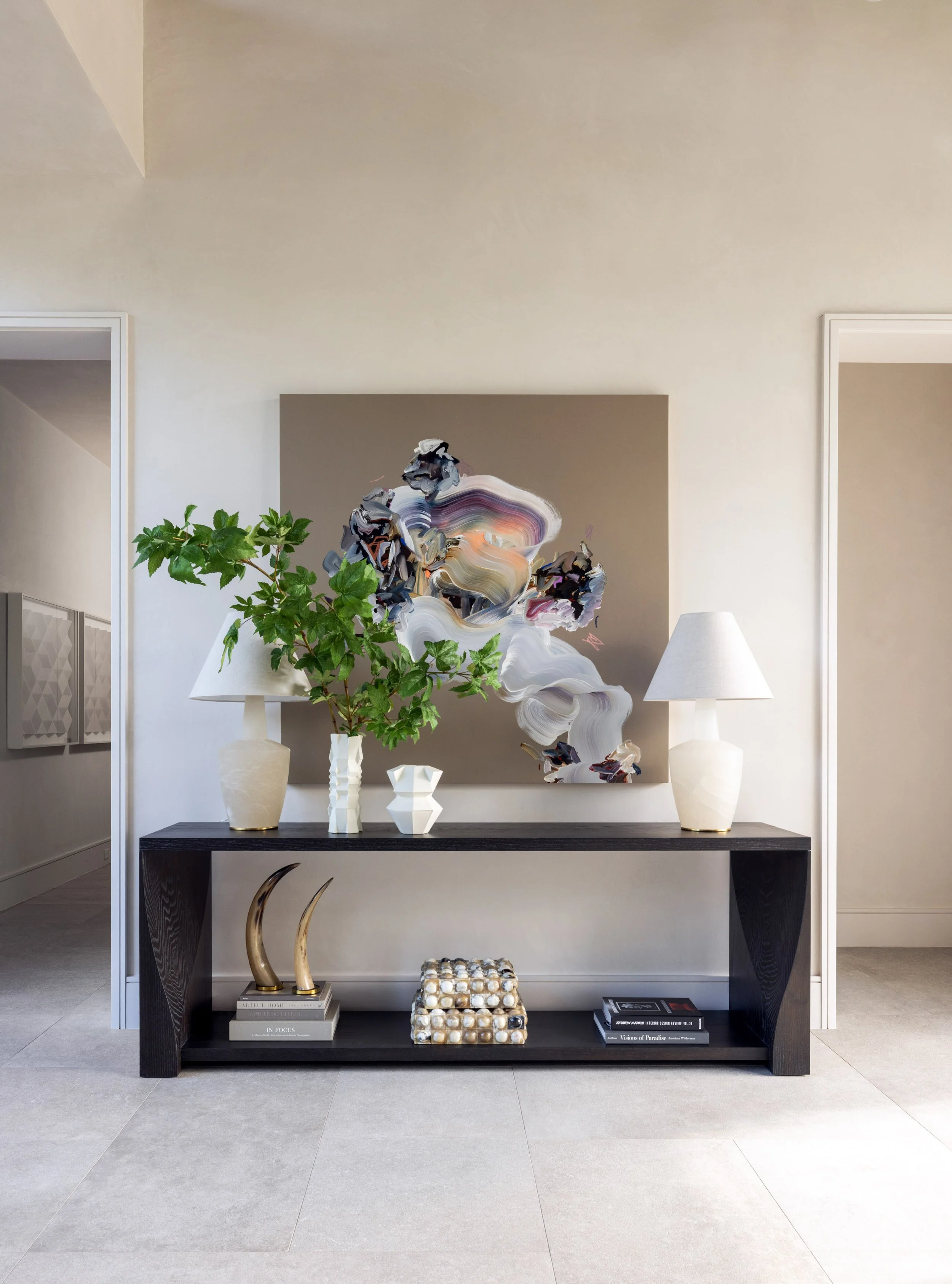 Benjamin Johnston Design entryway featuring dark console table with abstract artwork, white ceramic lamps, sculptural vases, brass horn accent, and large scale botanical arrangement.
