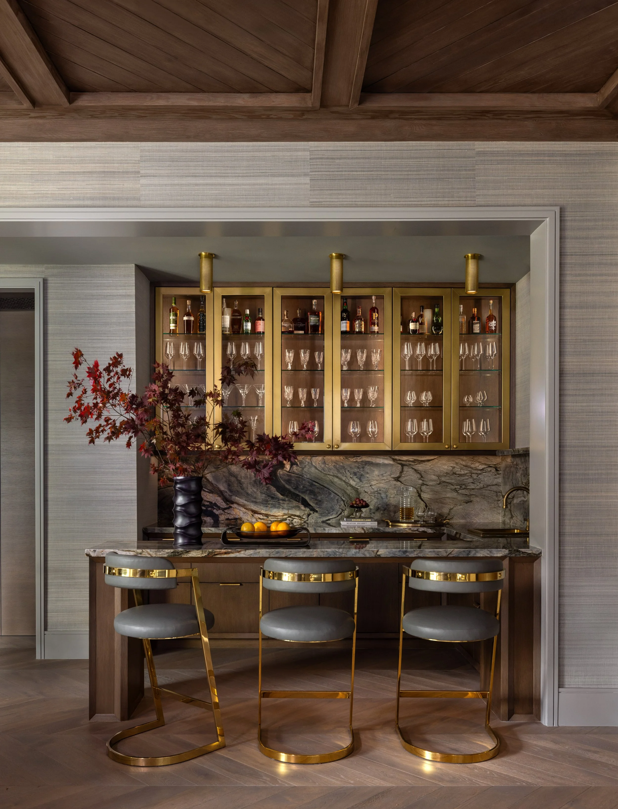 A home bar area with a marble countertop and gold accents. There are three modern bar stools with gold frames and gray cushions. Behind the bar, there is a gold cabinet with glass doors displaying various liquor bottles and glassware. A dark vase wit