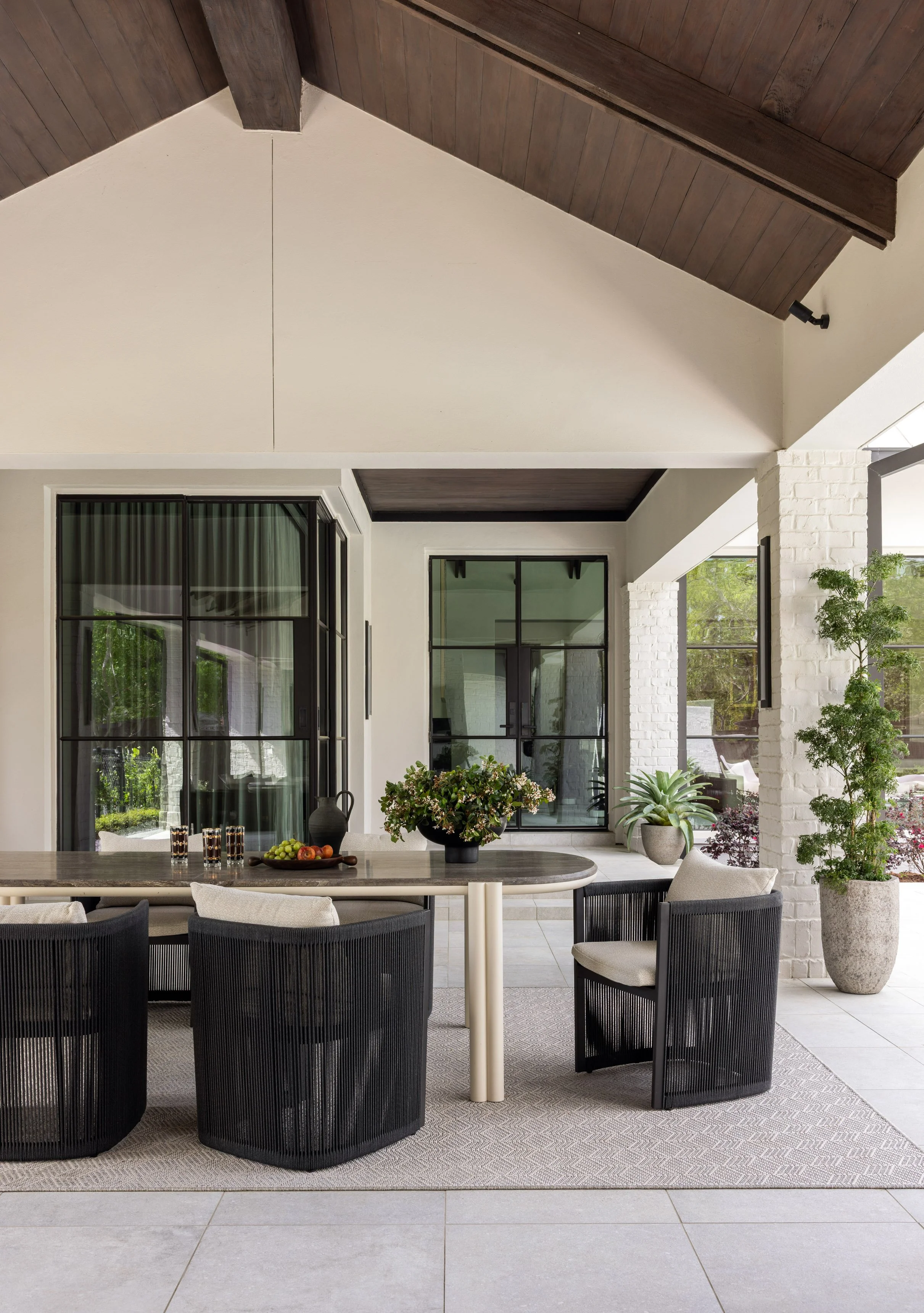 Benjamin Johnston Design Houston outdoor dining area featuring vaulted wood ceiling with exposed beams, woven black dining chairs, stone top dining table, floor-to-ceiling steel windows, and lush potted landscaping.