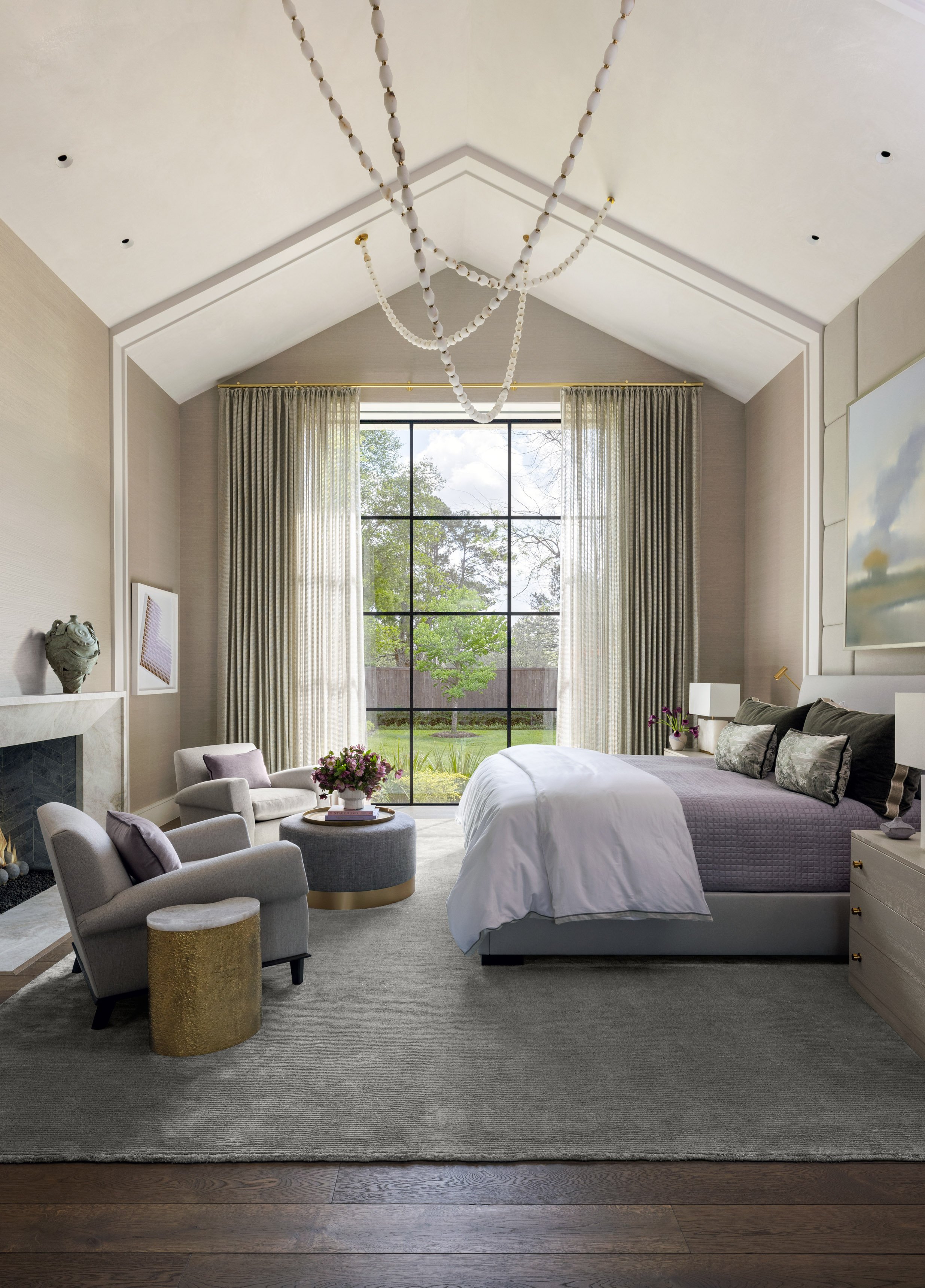 A spacious, modern bedroom with high vaulted ceiling, large window with sheer curtains, and a view of a green backyard. The room features a large bed with multiple pillows, a small white nightstand with a lamp, a comfortable lounge chair with a side 
