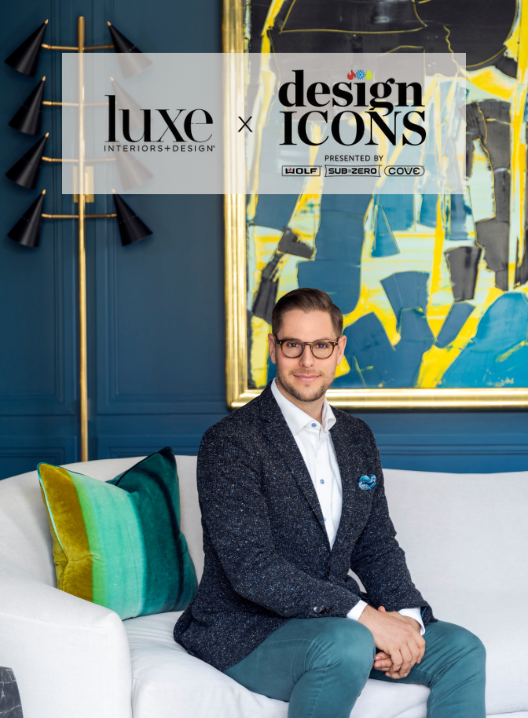 A man in a dark blazer, white shirt, and glasses sits on a white couch with colorful pillows in a stylish room with blue walls and modern wall art. A logo overlay reads "Luxe Interiors + Design" and "Design Icons."