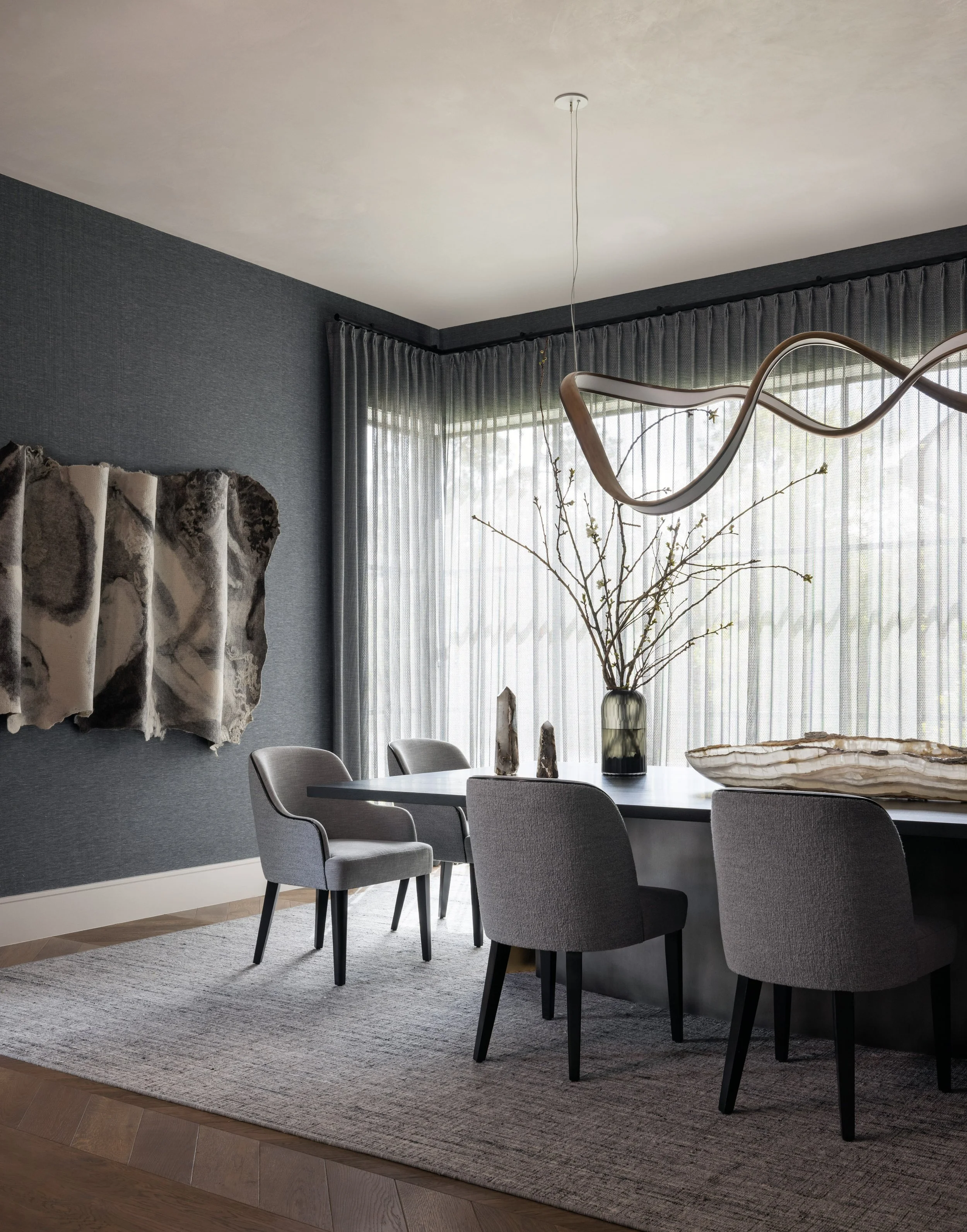 A modern dining room with a gray textured wall, a light wood table, six gray upholstered chairs, a gray area rug, decorative objects including a large glass vase with branches, and wall art on the wall.