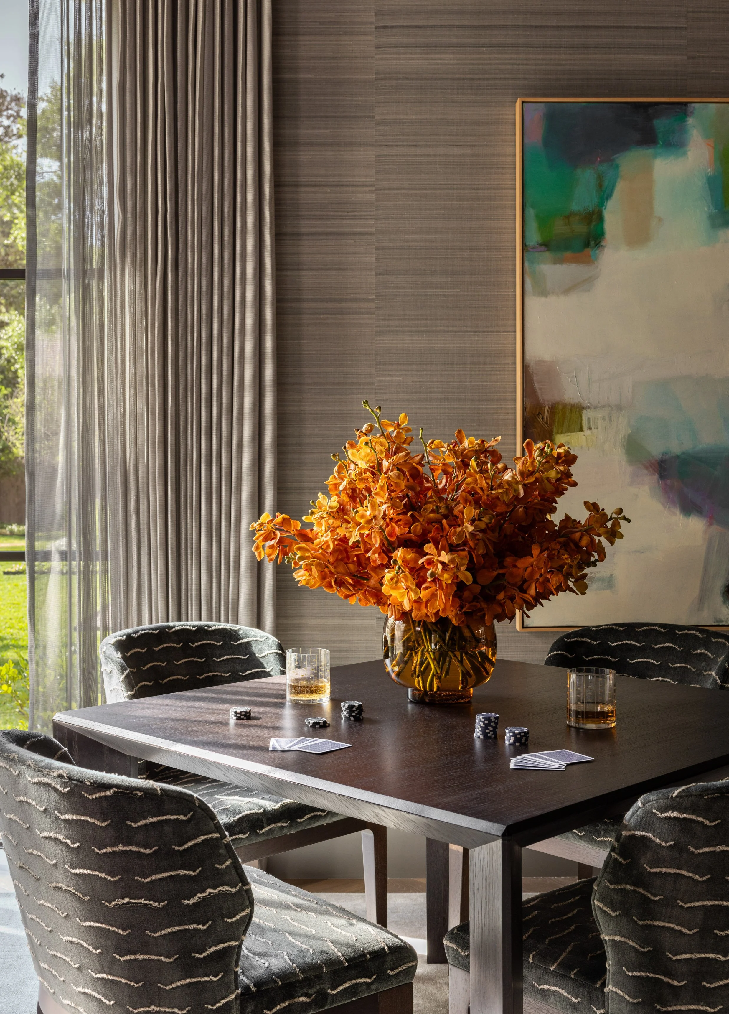 A dining room with a dark wooden table, six patterned chairs, and a large glass vase containing orange flowers. The table has some poker chips, playing cards, and glasses with a drink. There are beige curtains and a large abstract painting on the wal