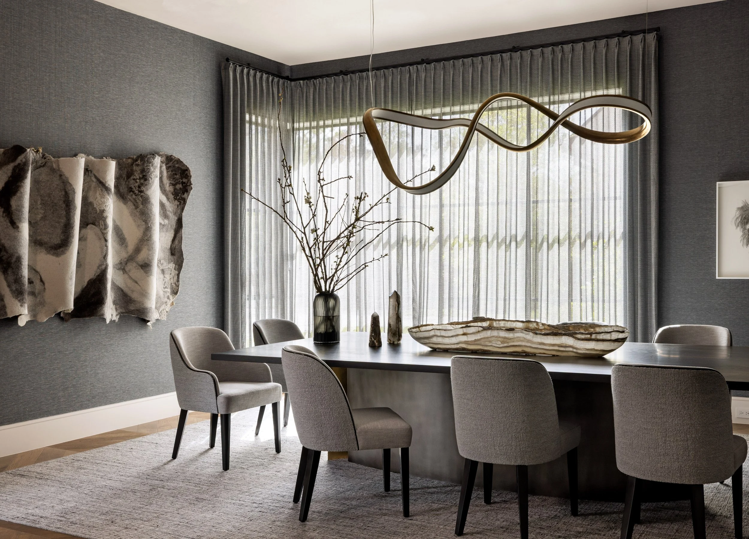 Modern dining room with grey textured walls, large window with grey curtains, and a rectangular dining table with grey upholstered chairs. A large decorative bowl and tall vases with branches are on the table. A unique, twisted chandelier hangs above