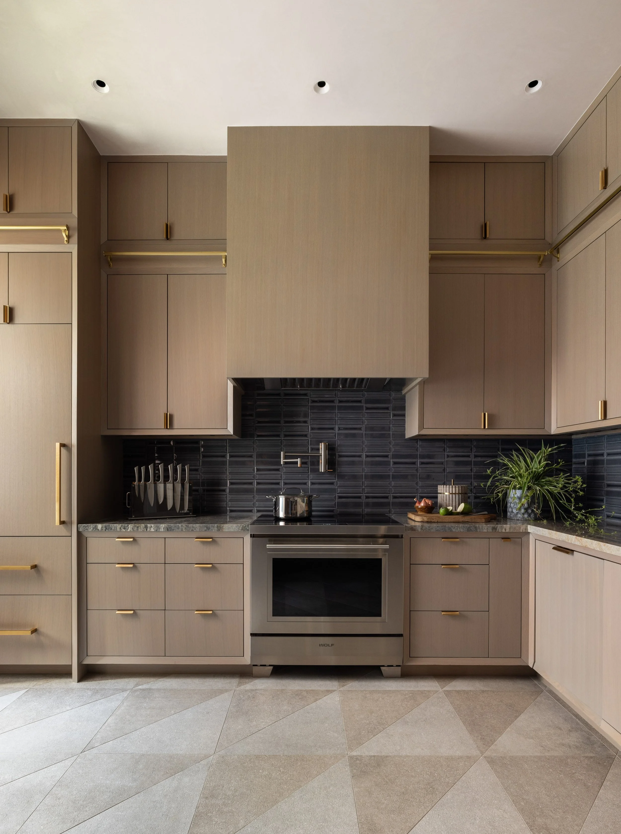 Modern kitchen with beige cabinets and gold handles, black tiled backsplash, stainless steel oven, and a countertop with a potted plant and a wooden tray with kitchen items.