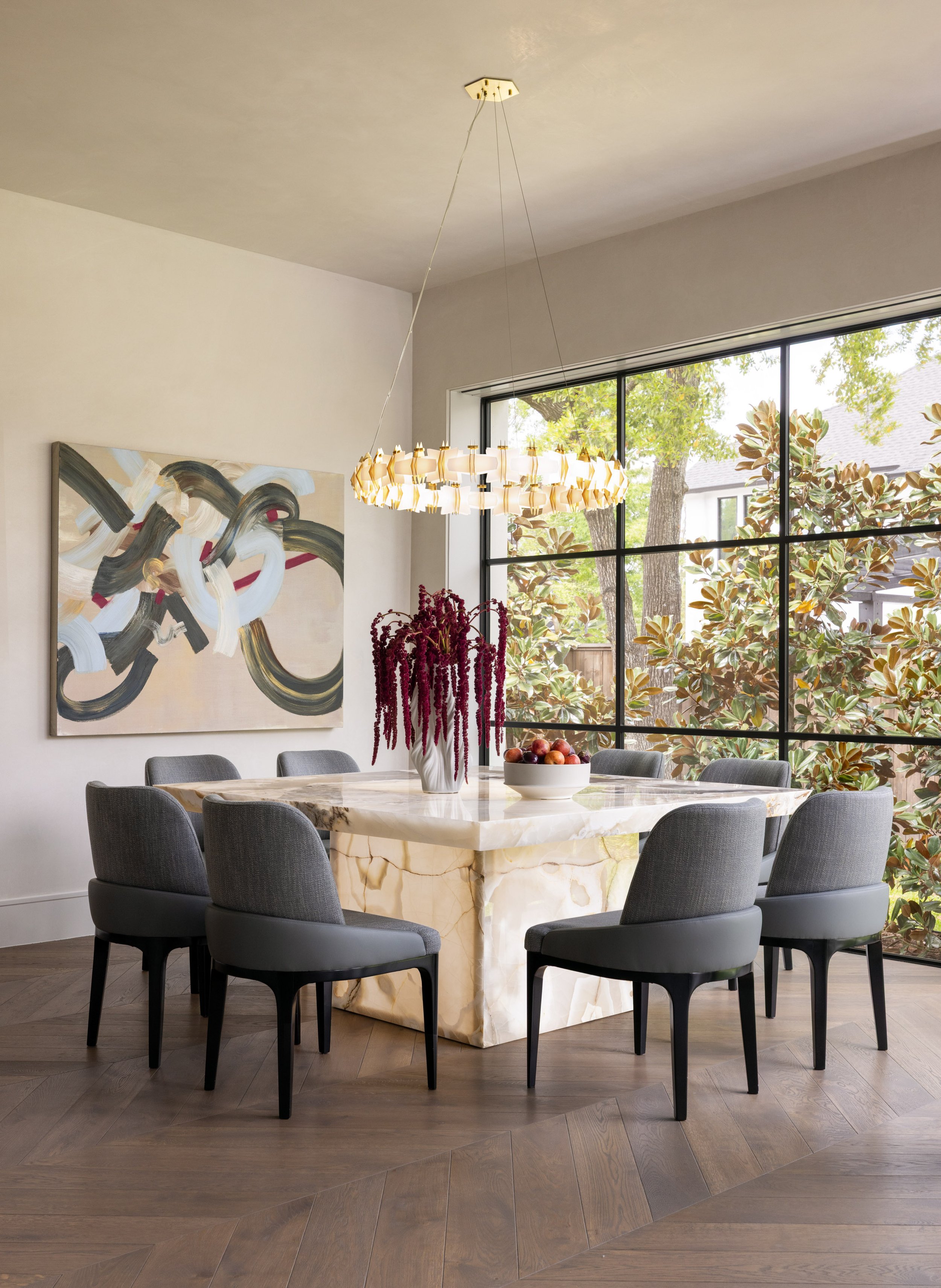 Modern dining room with a marble table, gray upholstered chairs, a vase with red flowers, abstract wall art, large window with greenery outside, and a hanging chandelier.