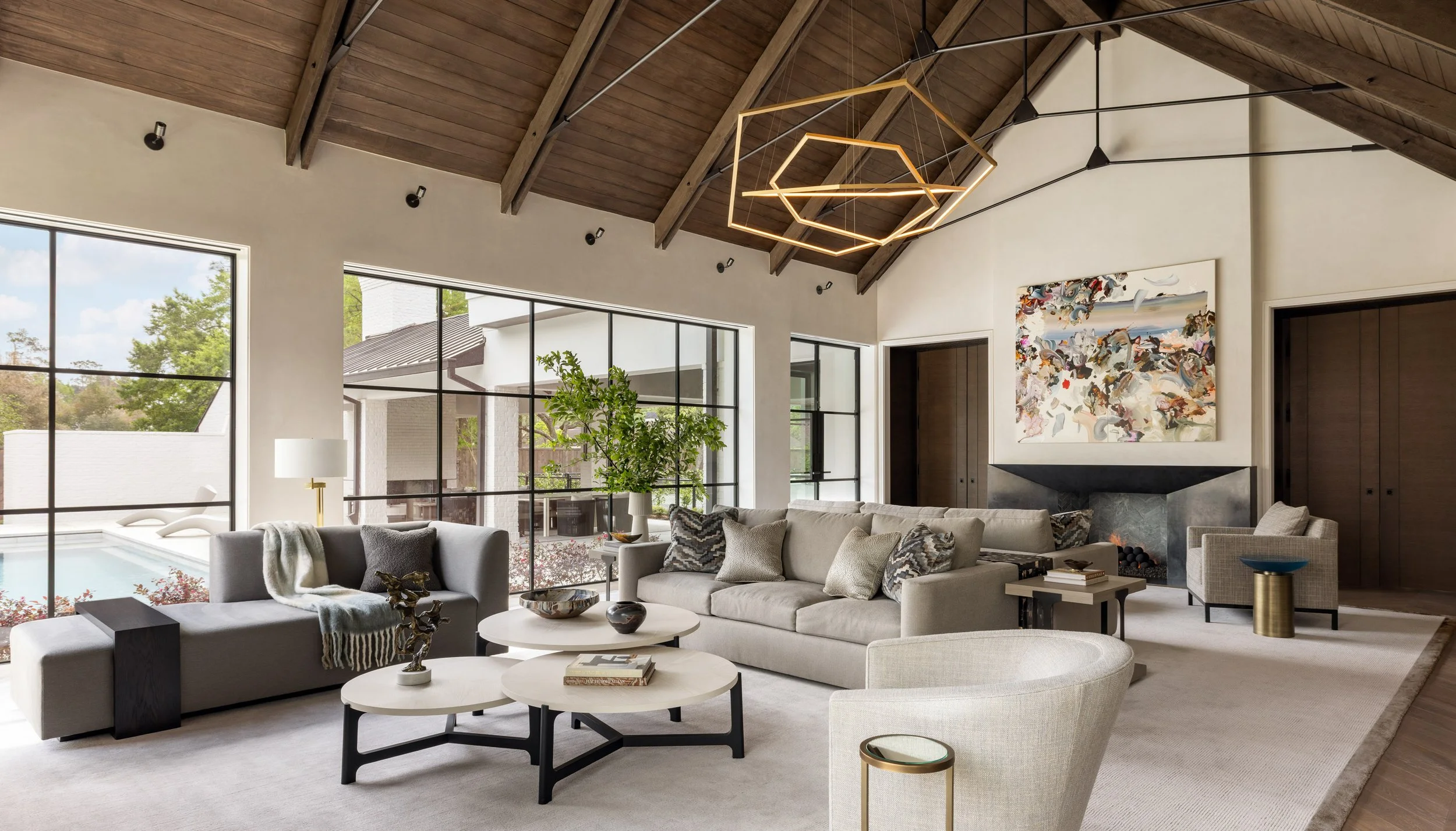 Benjamin Johnston Design Houston living room featuring soaring vaulted wood ceiling with exposed beams, geometric brass chandelier, floor-to-ceiling steel windows overlooking pool, neutral sectional sofas, abstract artwork, and stone fireplace.