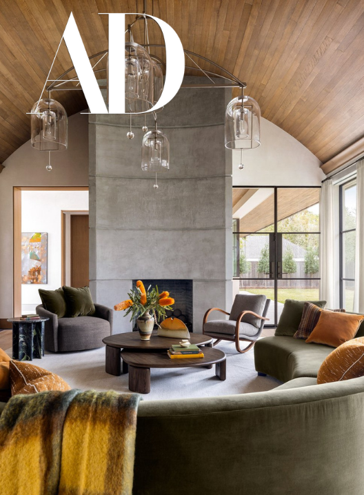 Modern living room with a high vaulted wooden ceiling, large windows, a concrete fireplace, and contemporary furniture including a sofa, armchairs, and a round coffee table with a floral arrangement.