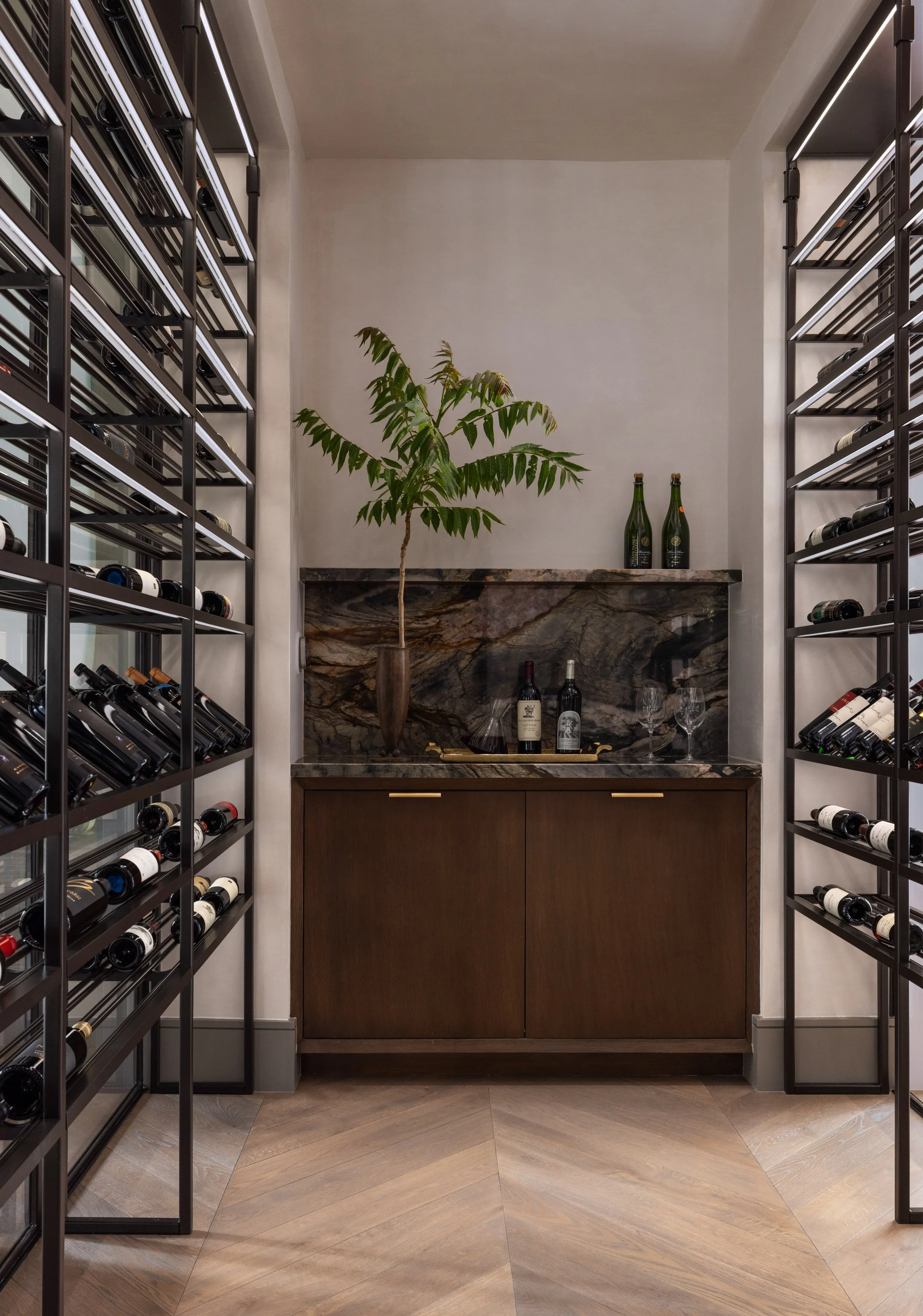 Benjamin Johnston Design wine room with illuminated floor to ceiling dark metal wine racks, walnut cabinet with brass hardware, veined marble backsplash and countertop, potted plant, wine bottles, and herringbone wood floor.