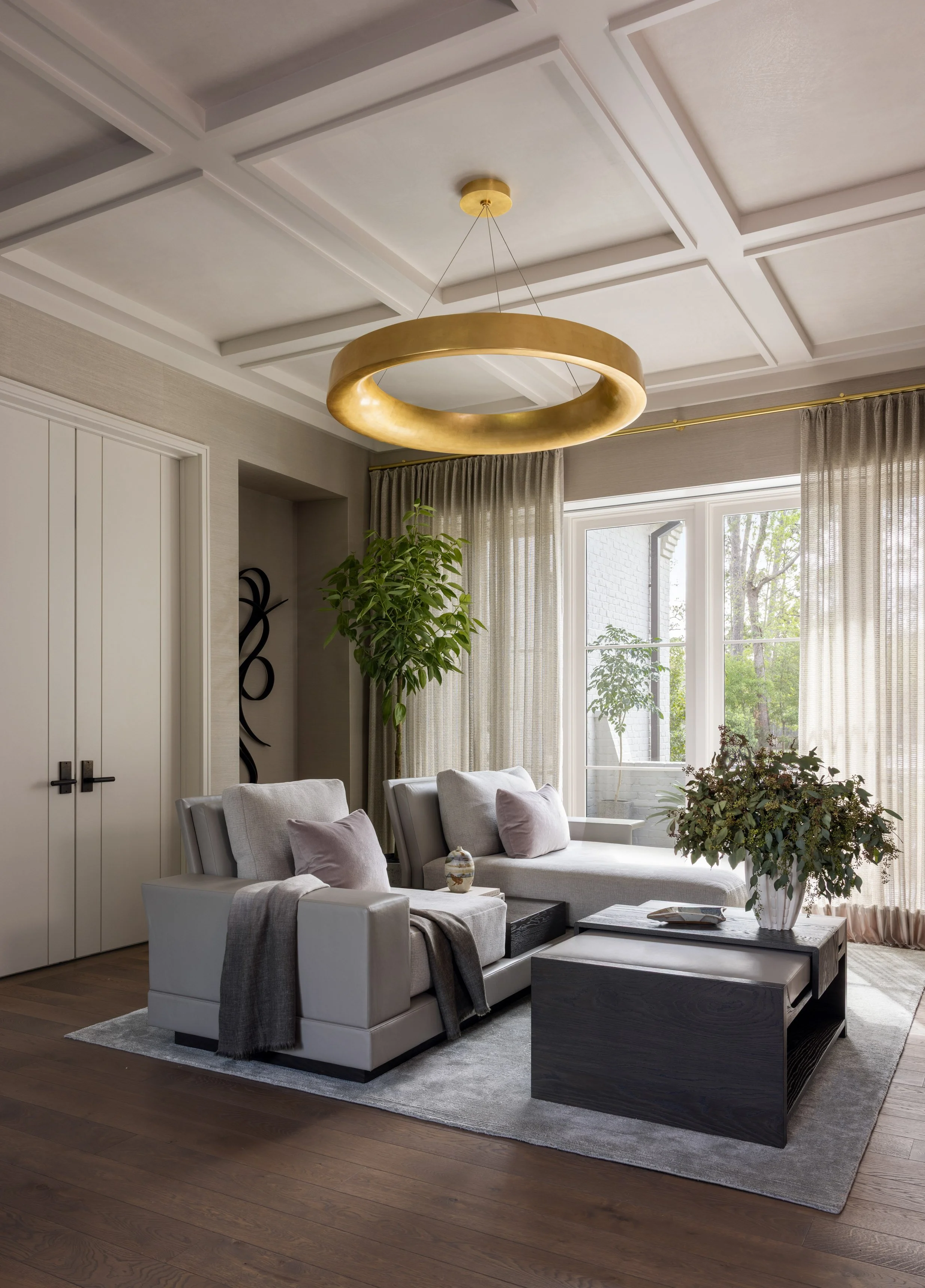 A modern living room with a coffered ceiling, a large gold ring chandelier, a large window with sheer curtains, white sofas with pillows, a black coffee table with books, a large potted plant, and wooden flooring.