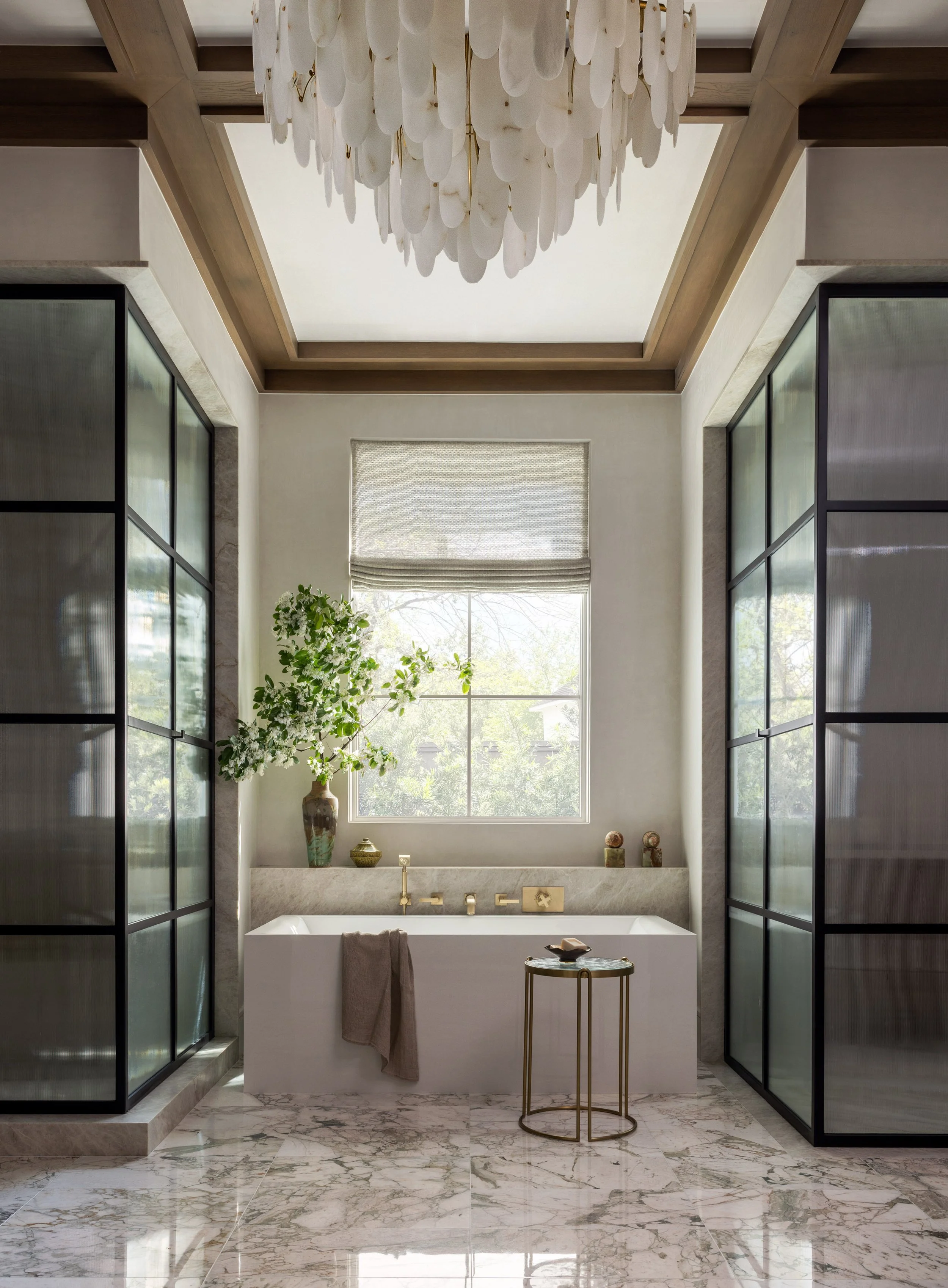 Benjamin Johnston Design primary bathroom featuring sculptural capiz shell chandelier, freestanding soaking tub with brass fixtures, marble tile floors, steel and glass shower enclosures, and botanical arrangement.