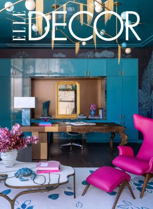 Interior decor with a blue wall and cabinetry, a vintage wooden desk, a pink chair and ottoman, a white coffee table with pink books, and gold and white hanging lighting fixtures.