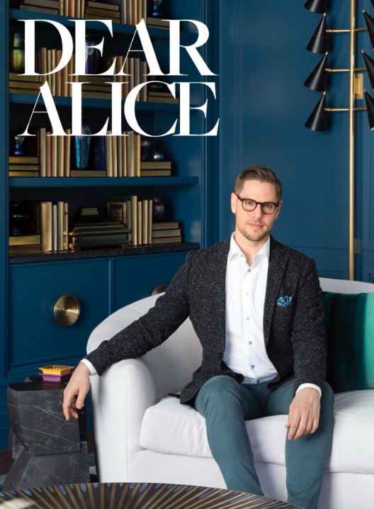 A man in glasses, a white shirt, a dark blazer, and teal pants sitting on a white sofa in a modern living room with dark teal walls and a bookshelf, next to the text "Dear Alice" in large white letters.
