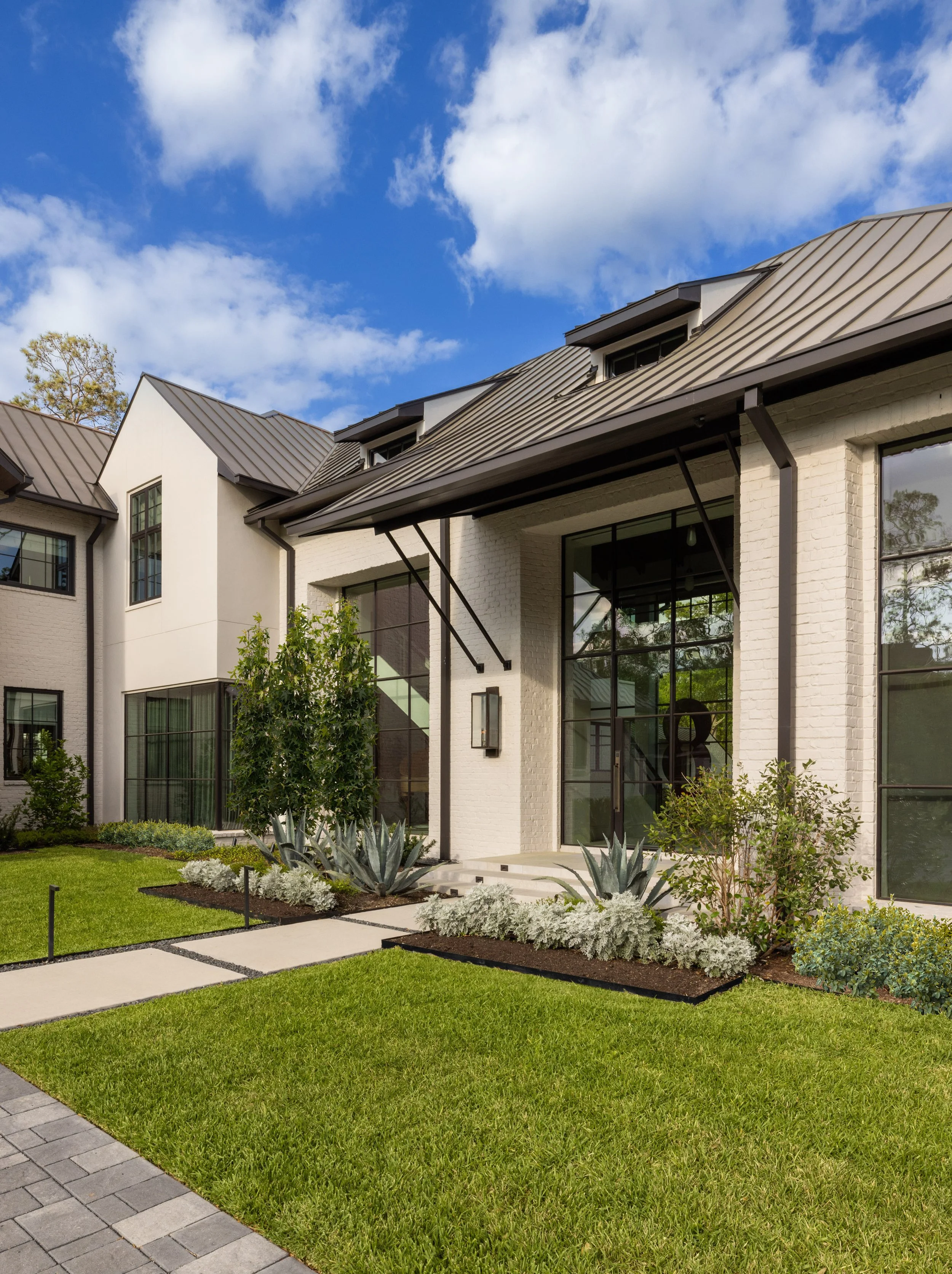 Benjamin Johnston Design residential architecture entry detail featuring white brick facade, standing seam metal roof, oversized steel entry doors, dramatic steel and glass window walls, and sculptural agave landscaping.