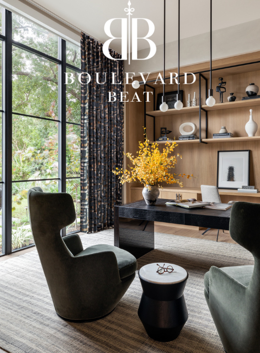 Interior of a modern living space with large windows, a black table with a yellow floral arrangement, two dark green chairs, a small black side table, wooden shelving with decor items, and patterned curtains.