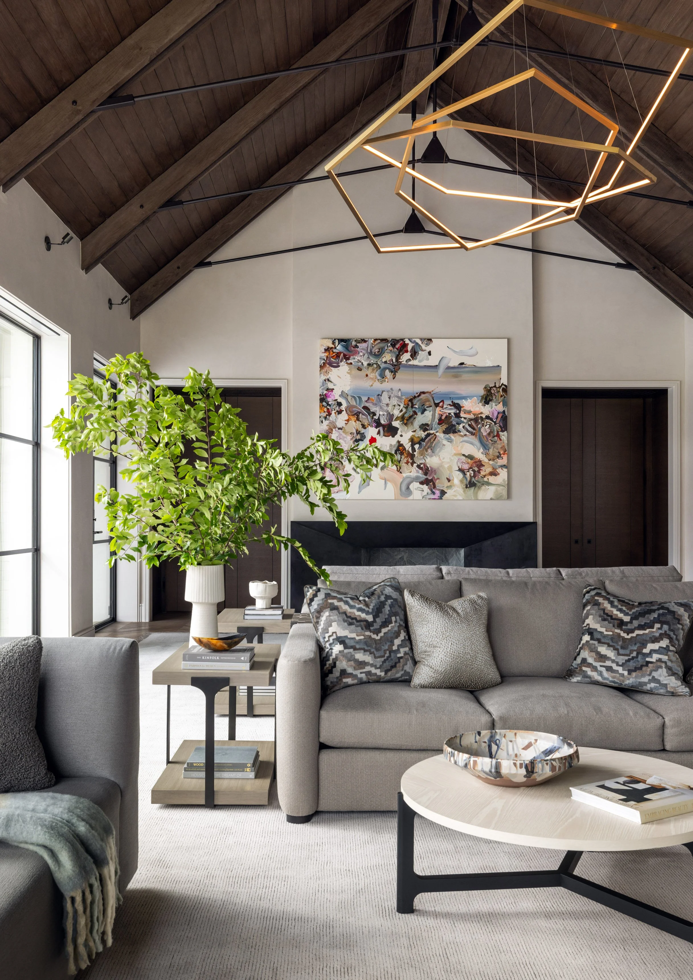 Benjamin Johnston Design Houston living room featuring vaulted wood ceiling with exposed beams, geometric brass chandelier, gray sectional sofa with patterned pillows, abstract artwork above stone fireplace, and large botanical arrangement.