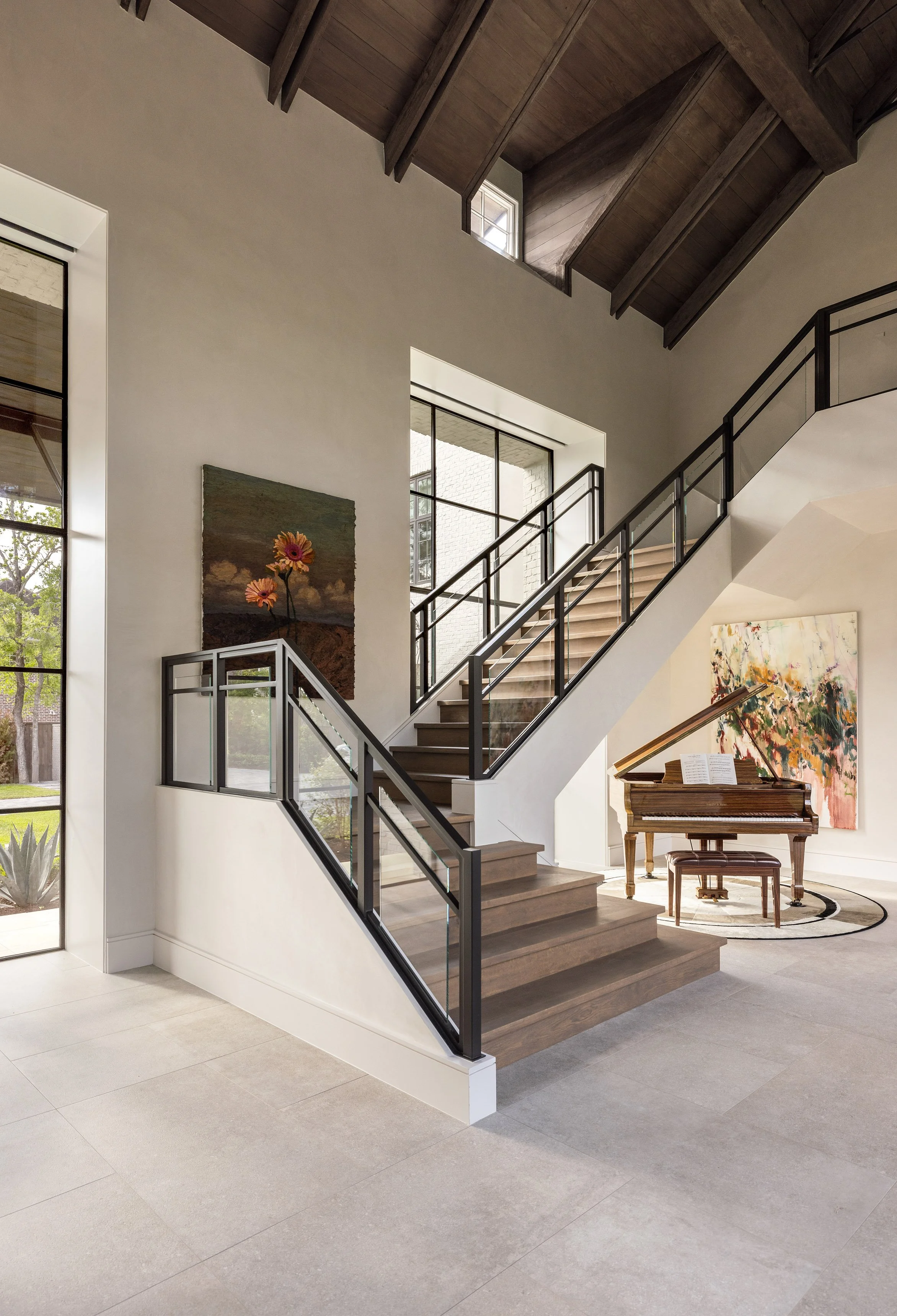 Benjamin Johnston Design entryway staircase featuring black steel and glass railing, vaulted ceiling with exposed wood beams, grand piano, colorful floral artwork, and floor-to-ceiling steel windows.