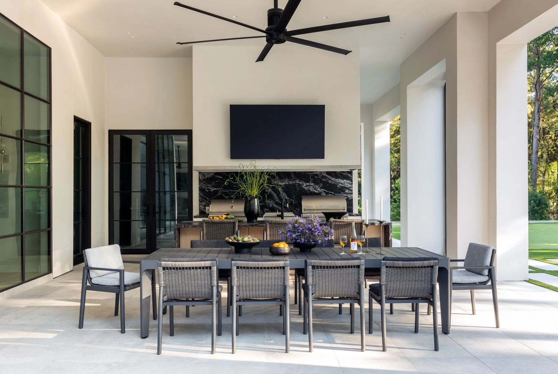 Benjamin Johnston Design Houston luxury residential interior design — covered outdoor dining area with built-in grill, black marble backsplash, mounted TV, woven dining chairs, and steel frame glass doors.