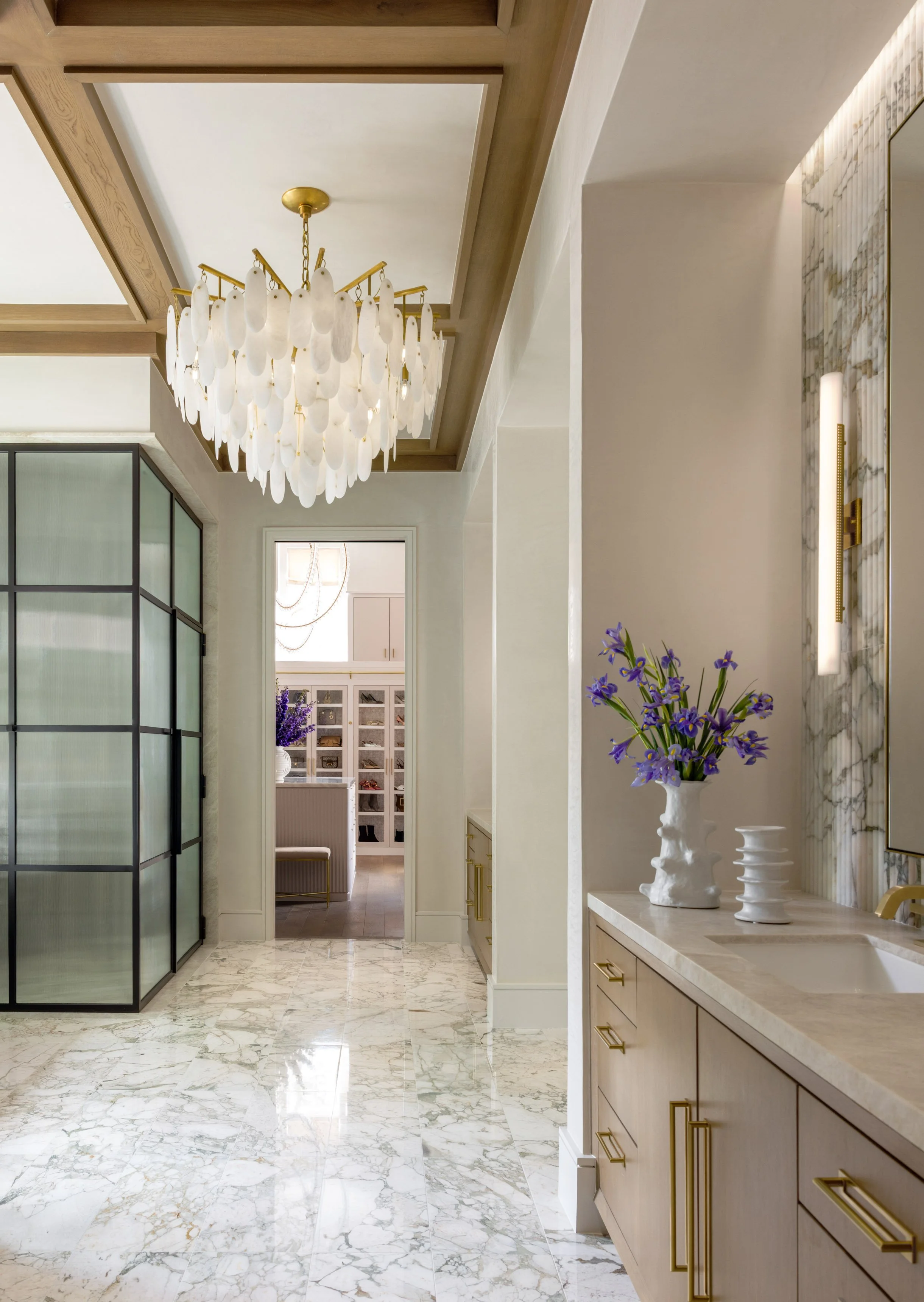 Interior of a luxurious home with marble floor, chandelier, decorative vases with purple flowers, and a marble wall with lighting.