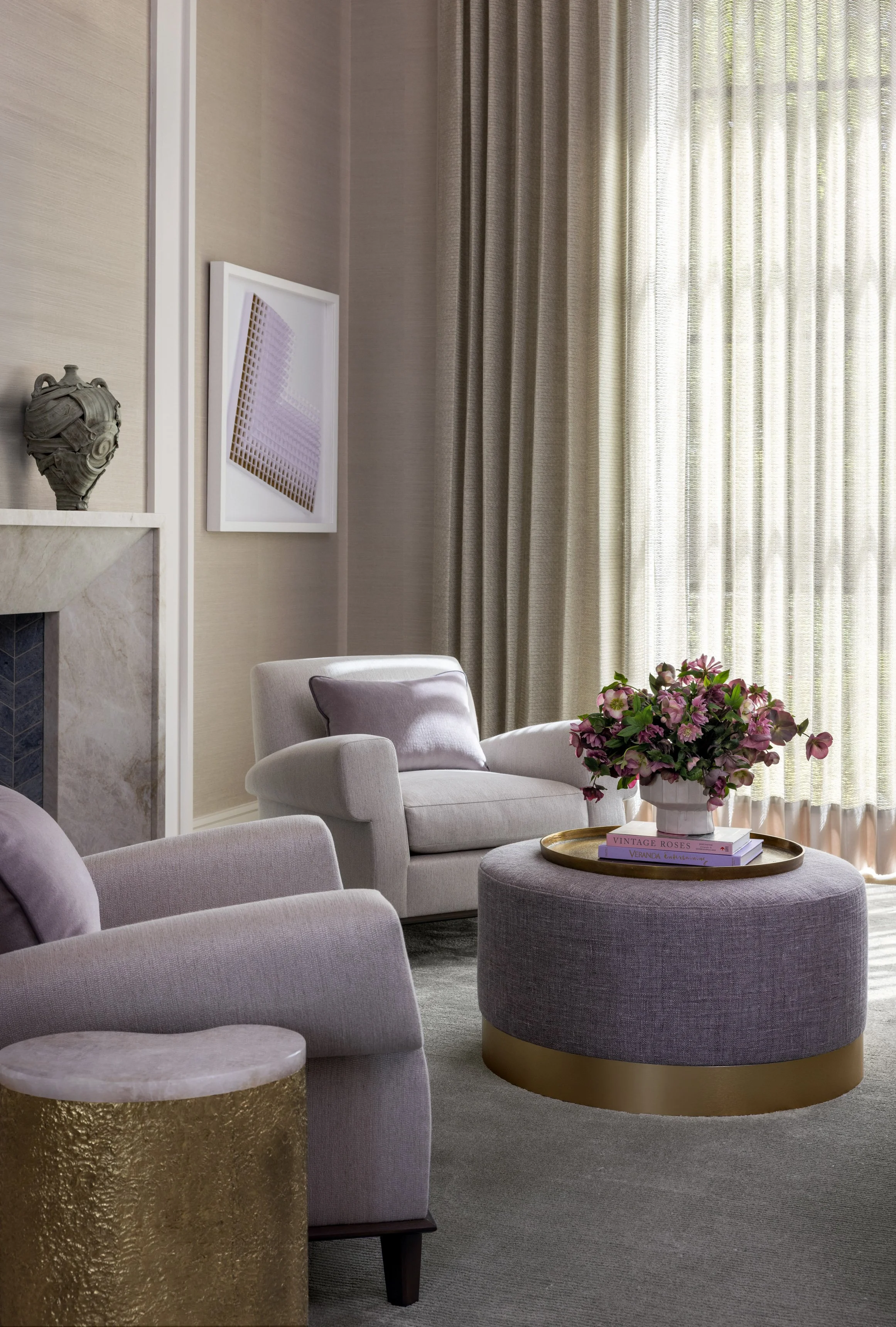 Benjamin Johnston Design primary bedroom sitting area featuring lavender lounge chairs, round upholstered ottoman with brass base, marble fireplace surround with chevron tile firebox, geometric abstract artwork, and sheer floor-to-ceiling drapery.