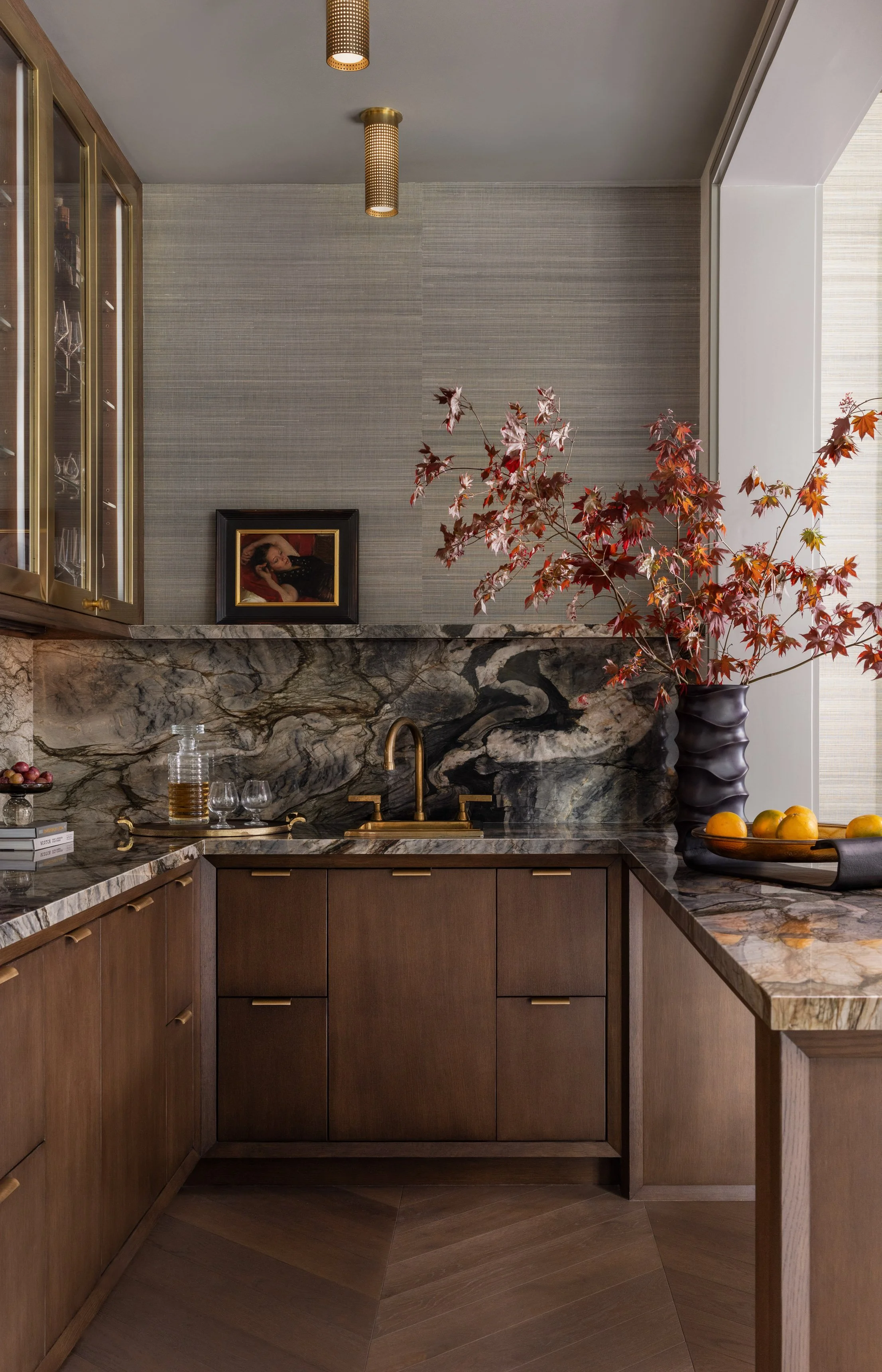 Benjamin Johnston Design wet bar with book matched marble backsplash, dark walnut cabinetry with brass hardware, brass sink and faucet, glass upper cabinets, grasscloth wallcovering, brass flush mount light, and red maple branches.