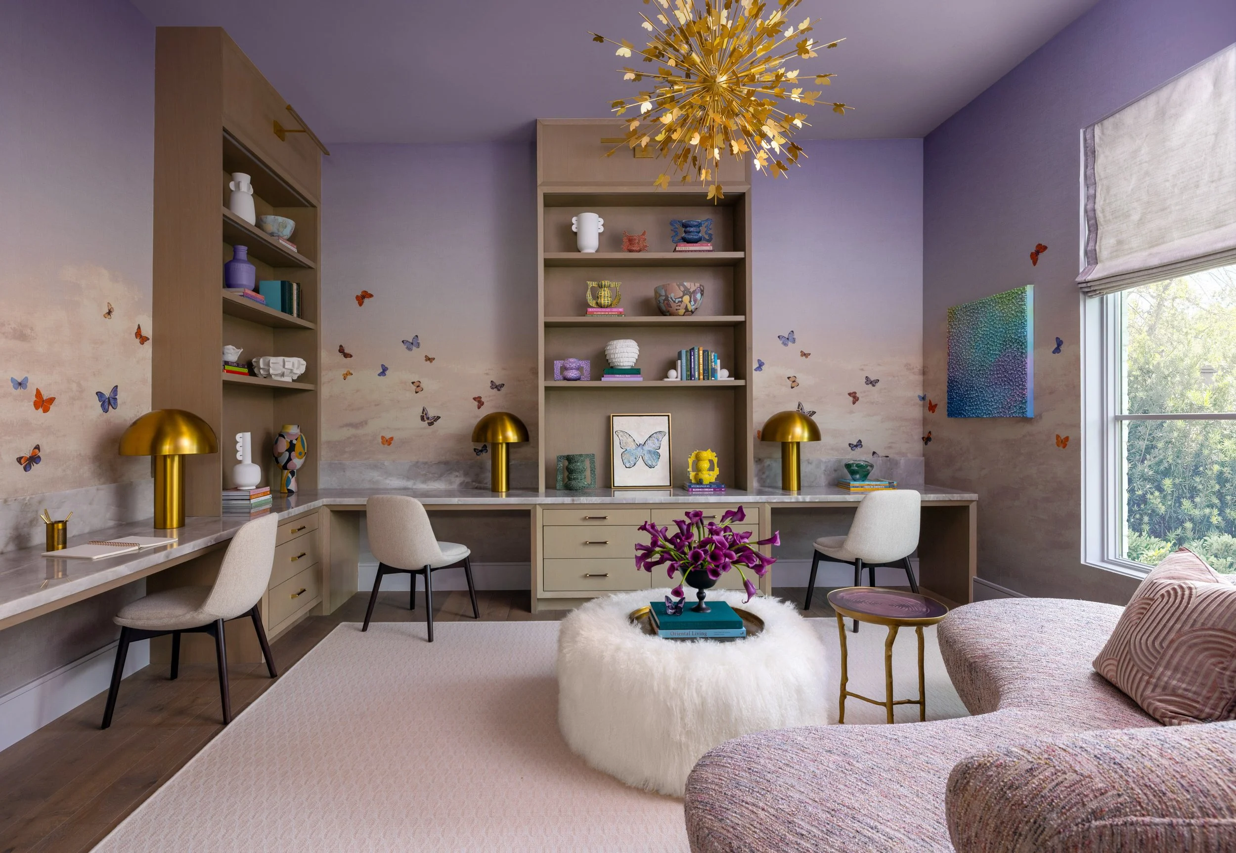 Benjamin Johnston Design children's study featuring lavender walls with butterfly wallpaper, gold sputnik chandelier, built-in oak shelving, dual marble desks with brass mushroom lamps, fluffy ottoman, and colorful curated accessories.